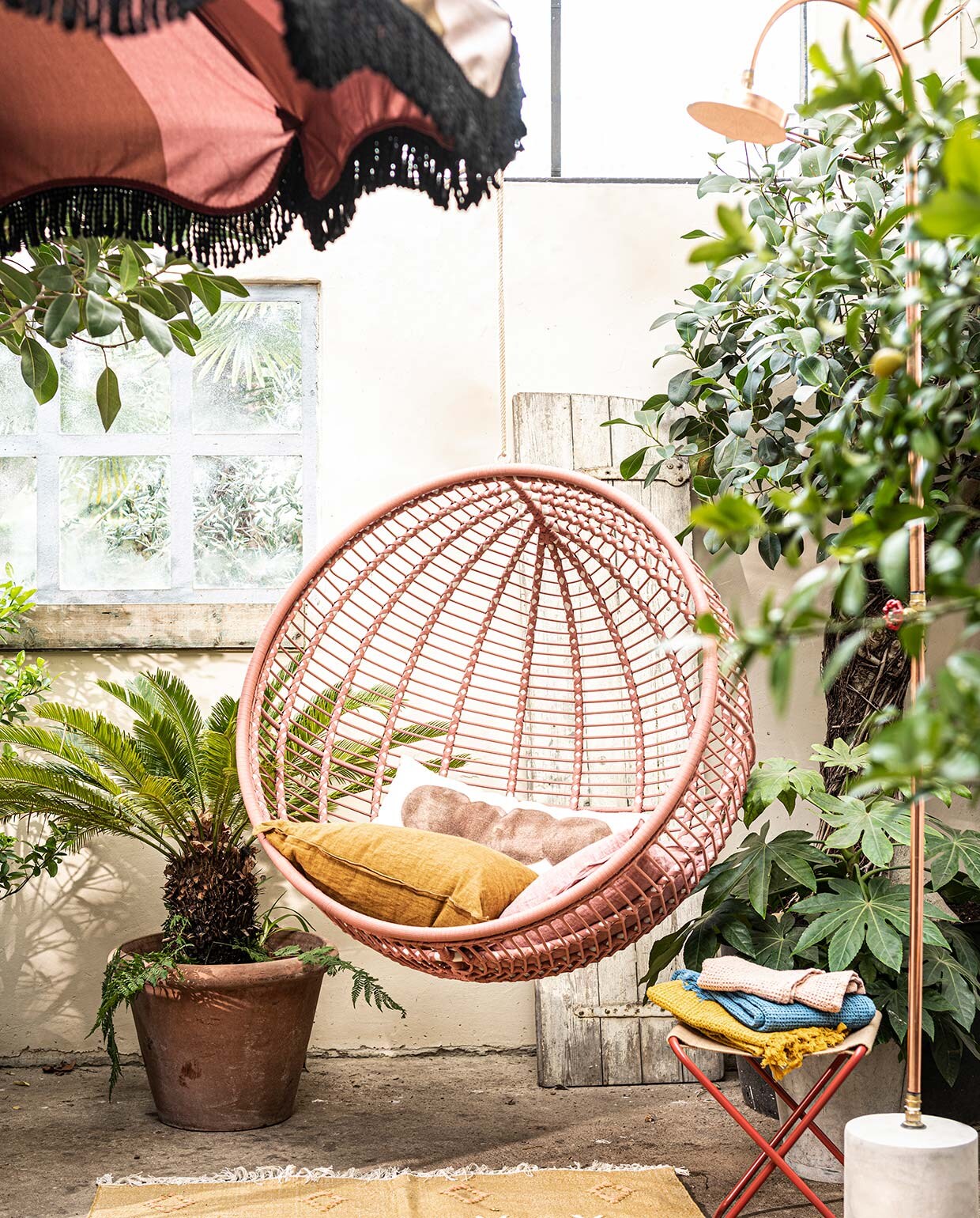 Stalen hangstoel voor buiten in zacht terra, 107×73 cm (Øxd)(kavehome.nl). Krukje Desert van Ferm Living (vtwonen.nl). Koperen buitendouche van Seletti (vtwonen.nl). Parasol van HKliving (vtwonen.nl). Langwerpig gestipt kussen (urbannatureculture.com ). Linnen kussens, wafeldoeken (bungalow.dk). Vloerkleed van Madam Stoltz (deens.nl). Plant en pot privébezit. vtwonen 07-2021 | styling Marianne Luning | fotografie Sjoerd Eickmans