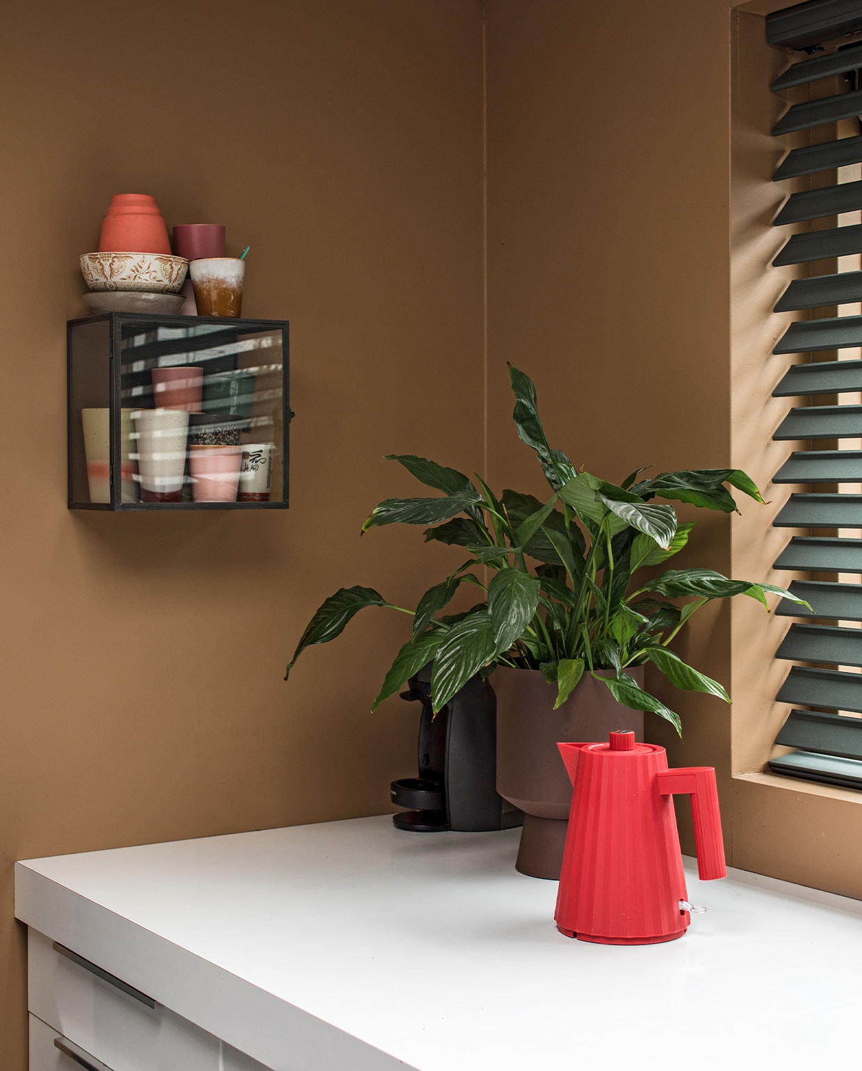 Muurverf Spiced Honey Flexa. Vitrinekast Ikea. Mokken HKliving, Hella Duijs, Nienke van de Pol. Servies kringloop. Koraalkleurig vaasje Hella Duys. Lepeltjes House of Oeli. Op aanrecht: bloempot Jysk, plant Ikea, rode theepot/waterkoker Alessi. Vlinderjaloezieën Bece raamdecoratie, Hubo. vtwonen 10-2021 | Productie en Fotografie Corine Hoge Bavel | Tekst Marion de Boer