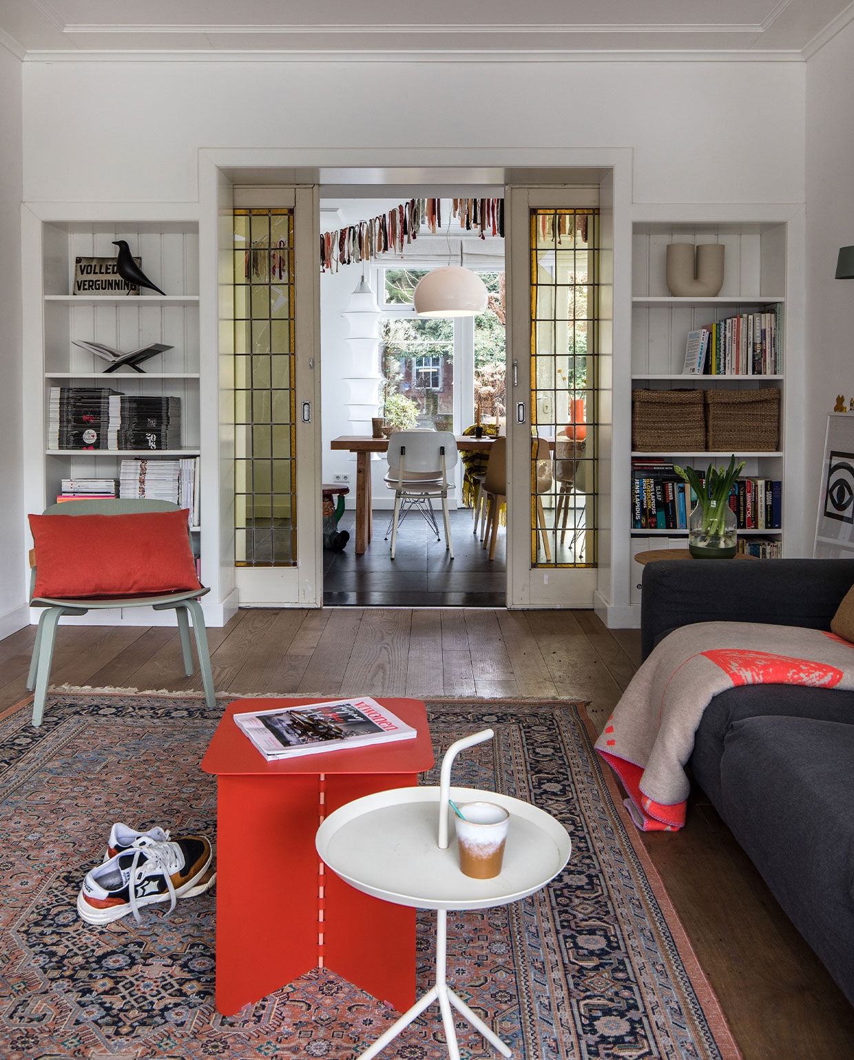 Vloer van Amerikaans eiken lag er al. Muurverf wit RAL 9016 Sikkens. Grijze bank gekocht tijdens een garagesale. Plaid op bank Home Sweet Home Adelheid Laden.Vloerkleed kringloop. Sneakers Atlantic Stars. Rode bijzettafel Puik Hinge. Witte bijzettafel Hay. Mok HKliving. Links in kast: drankvergunning uit café van vader, houten kraai Eames House Bird Vitra, boeken­standaard Mendo. Groene Lounge Chair Muuto. Rood kussen Kus-sen. Rechts in kast: vaas Kink Muuto, manden Ikea. Bruine bijzettafel Kwantum. Erop vaas Muuto. Schuifdeuren Marktplaats. vtwonen 10-2021 | Productie en Fotografie Corine Hoge Bavel | Tekst Marion de Boer