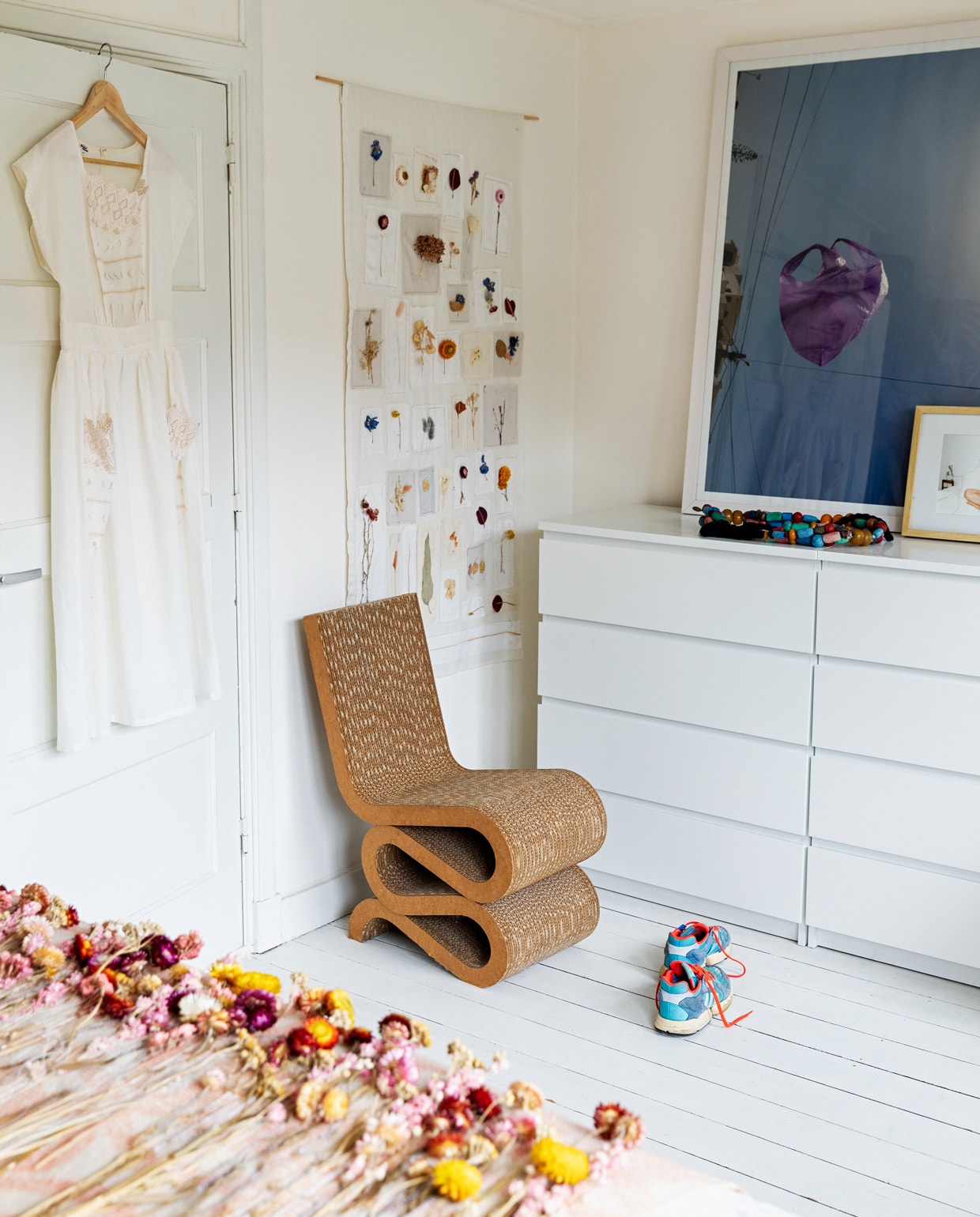 Wiggle Side Chair Vitra. Ladekast Ikea. Jurk vintage. Roze plaid met gedroogde bloemen en wanddoek met lapjes Loesje van Herp. Foto van paarse plastic zak Maarten Kuypers. Berber kralenketting op kast souvenir uit Essaouira (Marokko). vtwonen 13-2021 | Styling Ilona Jongepier | Fotografie Dana van Leeuwen | Haar &amp; make-up Djolien de Kreij | Tekst Eveline Stoel