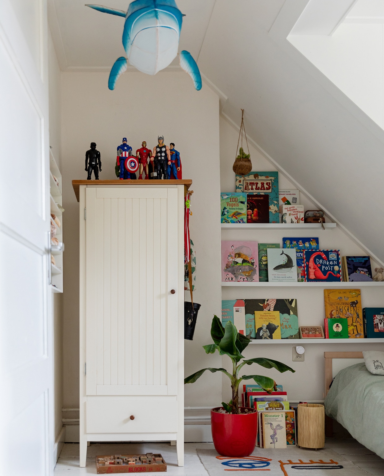 Papieren walvislamp Petit Pan. Witte kast, rode pot, houten letterdoos, letterbak aan de wand en Marokkaanse lamp op de grond kringloop. Tapijt Kwantum. Verzameling superhelden van de rommelmarkt. Bananenplant Intratuin. Bed en beddengoed Ikea. Grijs kussen met vogels Zilalila. Vintage verrekijker van opa. vtwonen 13-2021 | styling Ilona Jongepier | fotografie Dana van Leeuwen