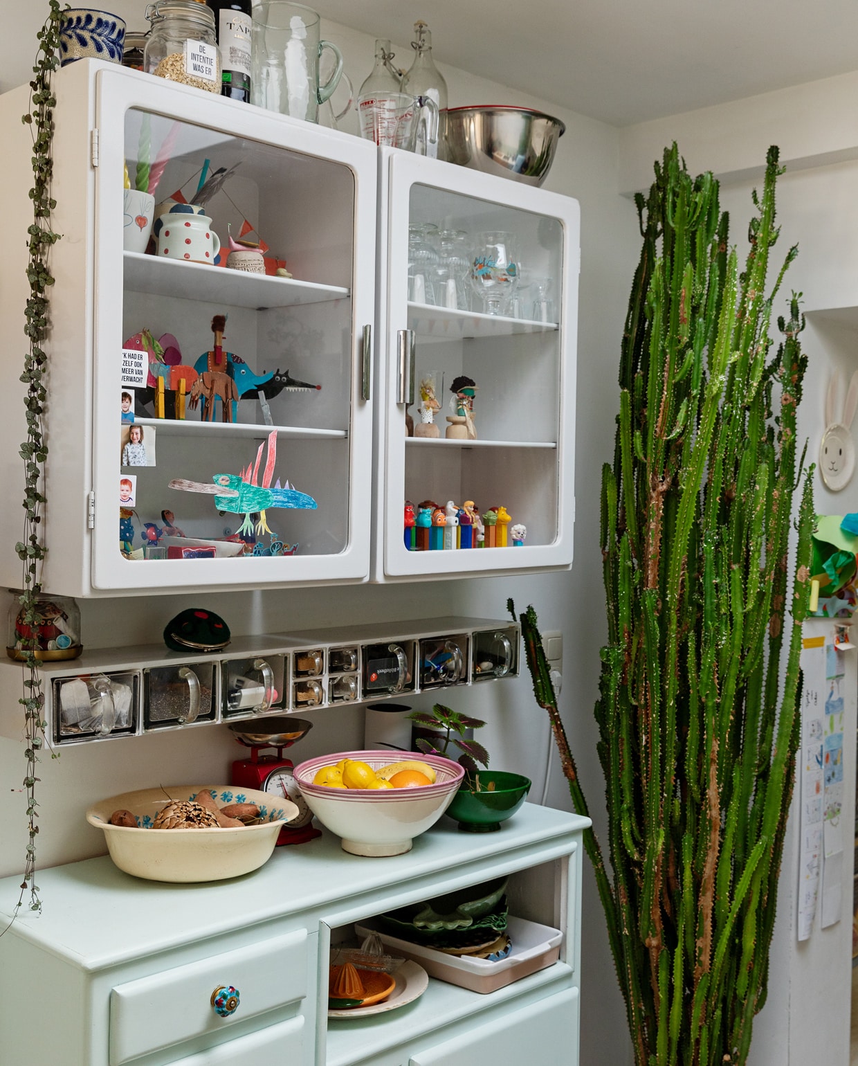 Vitrinekasten en kruidenrek Piet Zwart. Dressoir kringloop. Schalen en kommen Tine K Home en In-Petto. Weegschaal Dille &amp; Kamille. Mengkom rvs Duikelman. Grote cactus Marktplaats. Hangplant Ceropegia Intratuin. Pot kringloop. In de kast: verzameling Pez-snoephouders. Kartonnen dieren zelfgemaakt. Wijnglazen Iittala. Aardewerk meegenomen van diverse reizen. Tekeningen en rariteitenkabinet van Oskar. vtwonen 13-2021 | styling Ilona Jongepier | fotografie Dana van Leeuwen