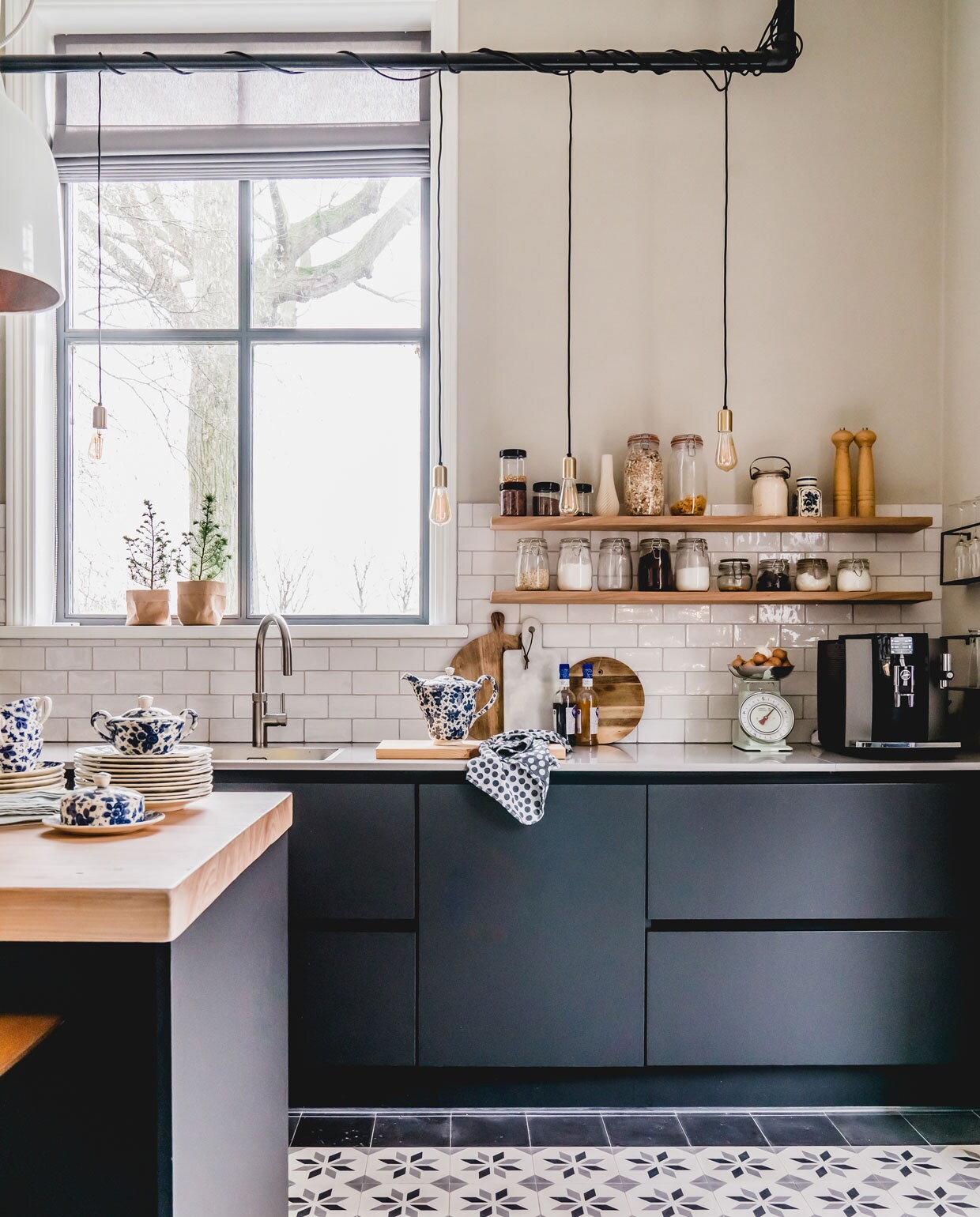 Ook de keuken kreeg nieuwe Portugese cementtegels van Floorz. De keuken is van Tulp. De reling met lampen maakten ze zelf van steigerpijp (steigerpijp.nl) en strijkijzersnoer (snoerboer.nl). De lamp boven de bar, waar ze graag aan ontbijten, is van Pols Potten. Het grijze vouwgordijn is van maatstudio.nl. Het blauwe servies komt uit de familie van Jankees, de voorraadpotten van Marjoleins opa. Plantenzakjes van Uashmama. vtwonen 12-2019 | Helenaveen binnenkijken oude pastorie keuken