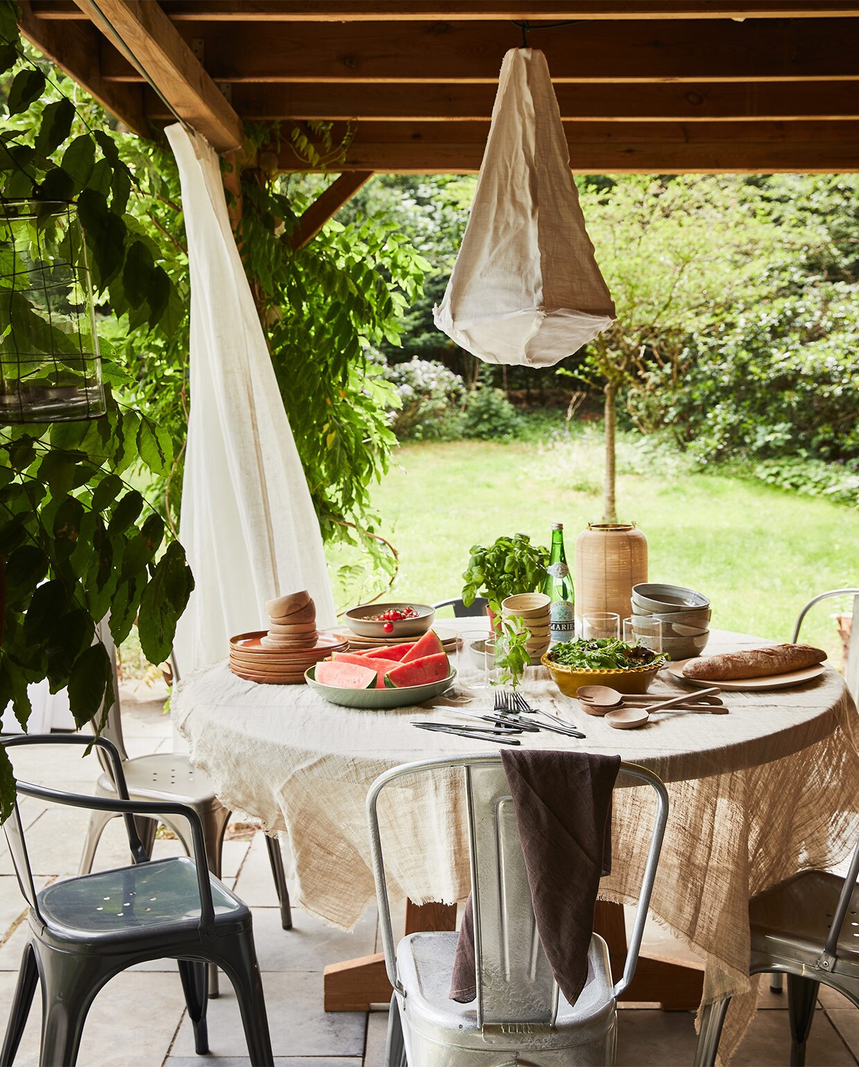 Teakhouten tafel, 140×75 cm (Øxh), kommen, diepe borden, platte borden, grote kommen met penseelstreek (hkliving.nl). Metalen stoelen (extravert.nl). Gordijnen Hilja, 145×300 cm (ikea.com). Hanglamp Torch, 40×64 cm, tafelkleed van HKliving 150×250 cm, bruine linnen theedoek van vtwonen (vtwonen.nl). Olijfkleurige schaal, walnoten saladeset (householdhardware.nl). Rest privébezit. vtwonen special tuin 03 2021 | styling Fietje Bruijn | fotografie Alexander van Berge