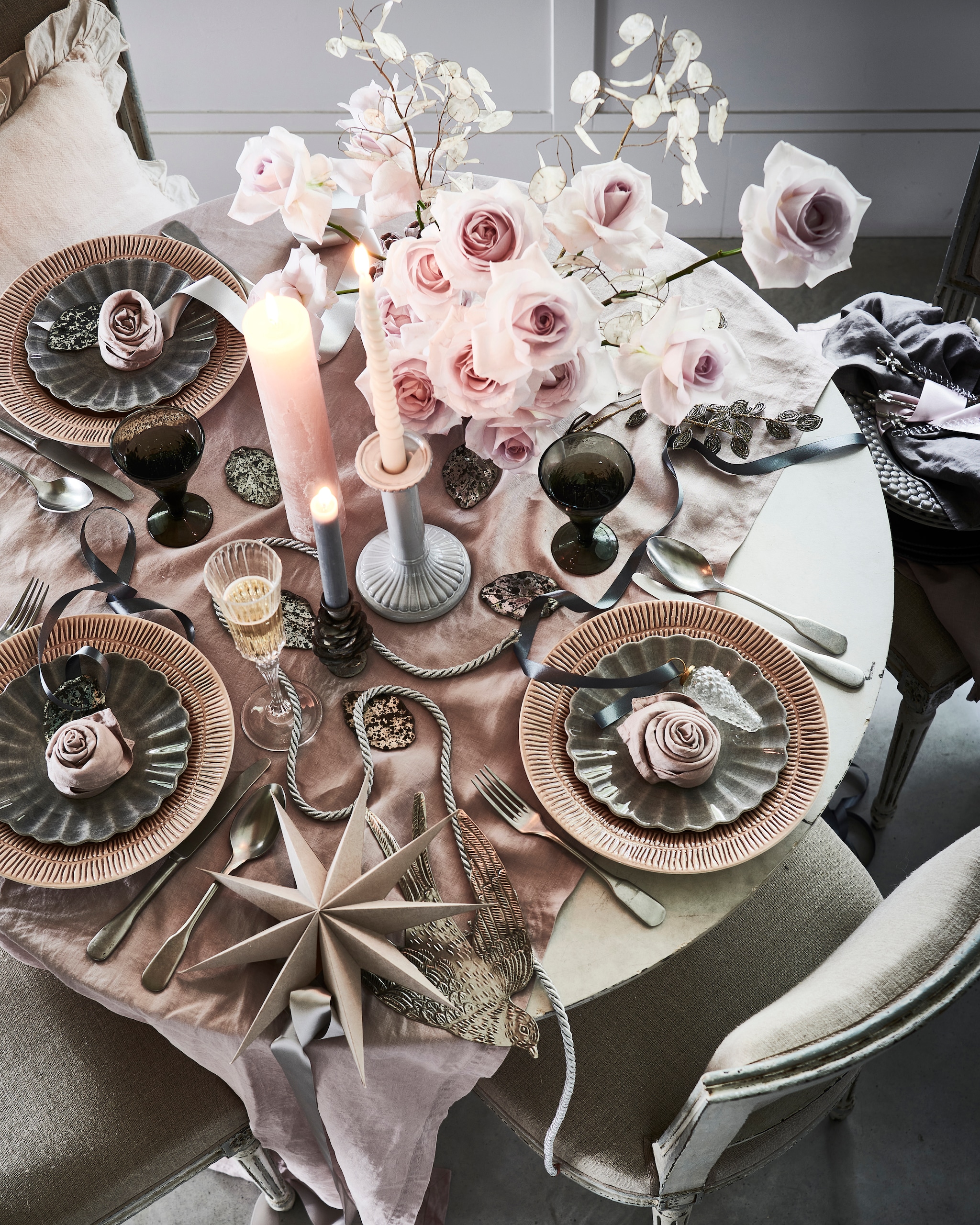 Bloemen, prijs op aanvraag (tuinvanjudith.wordpress.com). Tafel € 439,- (basiclabel.nl). Antieke stoelen, per stuk (pietjonker.nl). Linnen tafelkleed € 39,99, servetten, per set van 2 € 9,99, witte kandelaar € 17,99 (hm.com). Roze borden, per stuk € 29,50, grijze borden in bloemvorm, per stuk € 22,50, grijze borden met schulprand, per stuk € 30,95 (zwaanzinnig.shop). Roze kussen met volantrand € 22,99, champagneglas € 7,99 (zarahome.com). Zachtroze kaars € 14,- (allestedlund.com). Zinken kandelaar in vorm van dennenappel € 7,95, zilveren metalen zwaluw € 17,50, kerstdecoratie van kralen in vorm van blad, kerstbal dennenappel € 4,95 (weldaad.com). Grijze wijnglazen, per stuk (anna-nina.nl). Grijze papieren ster € 8,- (bungalow.dk). Theelepels van gedraaid glas, per stuk € 3,99, zilveren theelepels met vogel, per stuk € 4,99 (sissy-boy.nl). Interieurontwerp Piet-Jan van den Kommer (vandenkommer.nl). Overig privébezit. Tutorials over een roos maken van een servet staan op youtube onder servetvouwen.nl. vtwonen 12-2021 | styling Danielle Verheul | fotografie Sjoerd Eickmans | tuin van Judith (groenstylist) | assistentie Niels Kist, Kula van den Broek