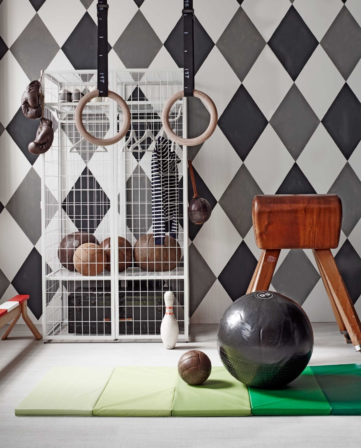 Kast Ikea PS 2014, gymmat Plufsig, evenwichtsbank Ikea PS 2014, set kledinghangers Bumerang (Ikea). Oude gymbok (Brût Amsterdam). Houten gymringen MP122 en gymbal van Adidas Ø 65 cm (www.muscle-power.nl). Oude leren gymballen en leren bokshandschoen (Spoor 38). Leren trainingsbal met leren lus en kegel (Neef Louis). Zwarte kist in kast (Combitex). Het dessin is geschilderd in muurverf grijs Plummett 272, zwart Off-Black 57, wit Strong White 2001 (Farrow &amp; Ball). Gestreept vest (Petit Bateau). Gympen New Balance (Maison NL). vtwonen 07-2014 | Fotografie Alexander van Berge | Styling Cleo Scheulderman