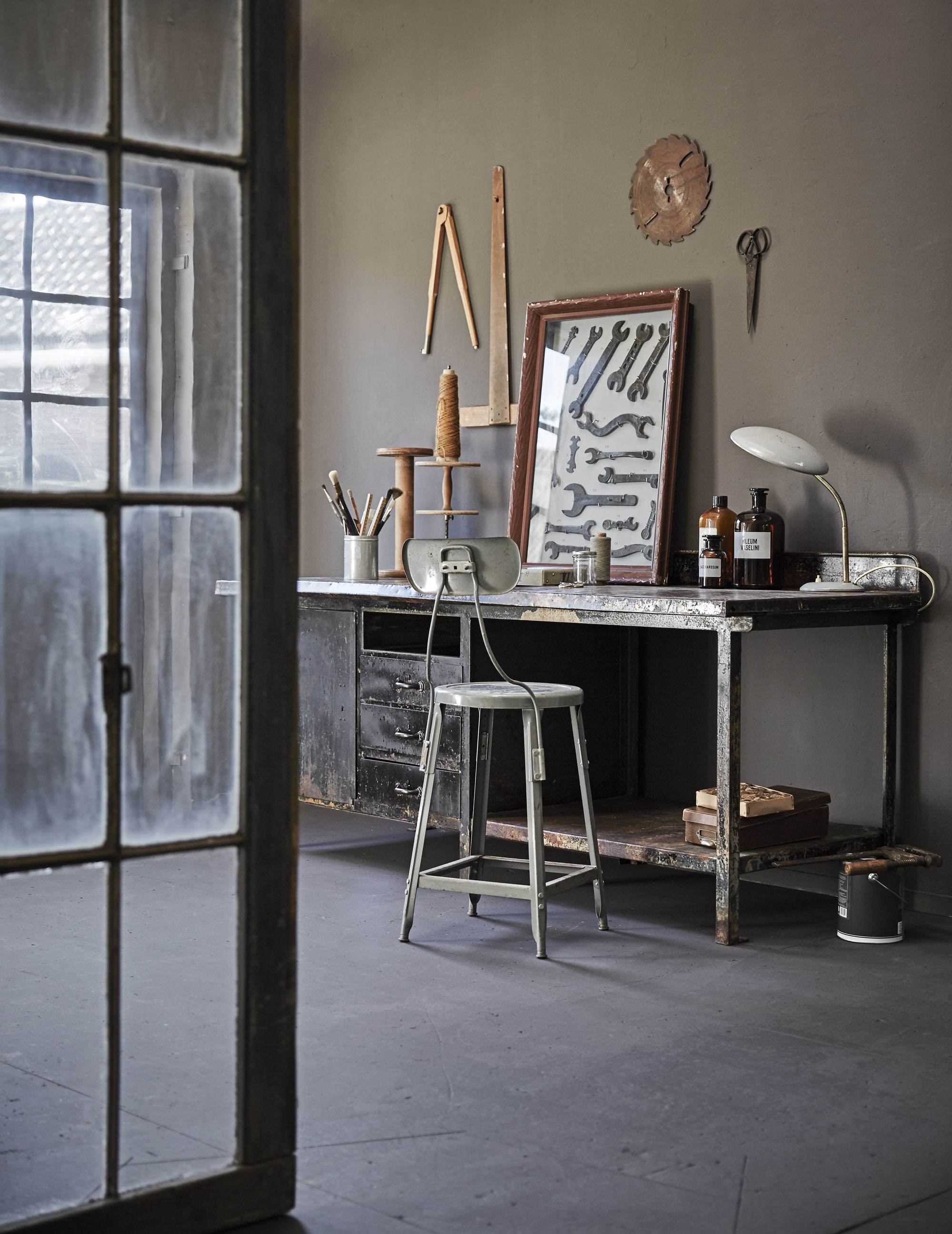 Fabrieksdeur met roedeverdeling (Combitex). Werktafel Stochov 75x197x80 cm (Brût Amsterdam). Werkplaatskruk (Spoor 38). Op het bureau: blik met kwasten, houten klos, garenhouder, ingelijste sleutels, bureaulamp (Spoor 38), apothekersflessen, schaar (Brût Amsterdam), doosje met garen (Combitex). Onder het bureau: bruine doos (Spoor 38). Aan de muur: passer (Six and Sons), liniaal (Neef Louis). Het overige is privébezit. Muurverf Mud Grey van vtwonen (vtwonen.nl).