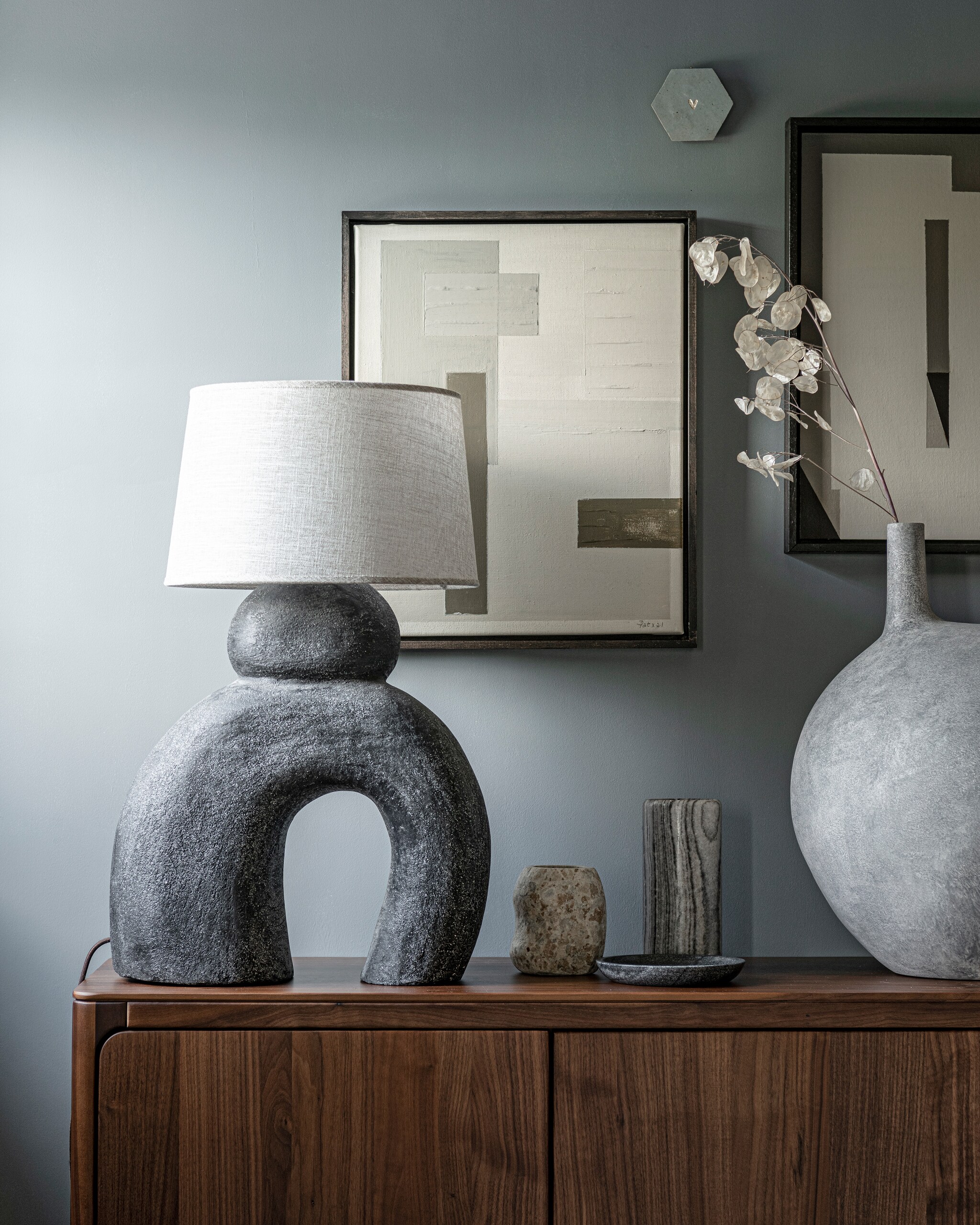 Kleur op wand Blue Stone van Pure &amp; Original 1 liter € 40,75 (pure-original.nl). Lamp Sur € 449,95 (lanena-home.com). Kunstwerken, prijs op aanvraag (dailypat.com). Handgemaakte tegel met gegraveerd hart € 23,95 (tegeltjetegeltjeaandewand.nl). Lichtgrijze vaas Submarine, big € 349,- (101cph.com). Marmeren vaas € 29,99 (hm.com). Stenen vaas € 45,99, stenen schaal € 22,99 (zarahome.com). Walnoothouten dressoir Rikke € 2960,- (demachinekamer.nl). vtwonen 11-2021 | Styling Kim van Rossenberg | Fotografie Sjoerd Eickmans | Assistentie Sanne van der Beek en Meike Linthorst