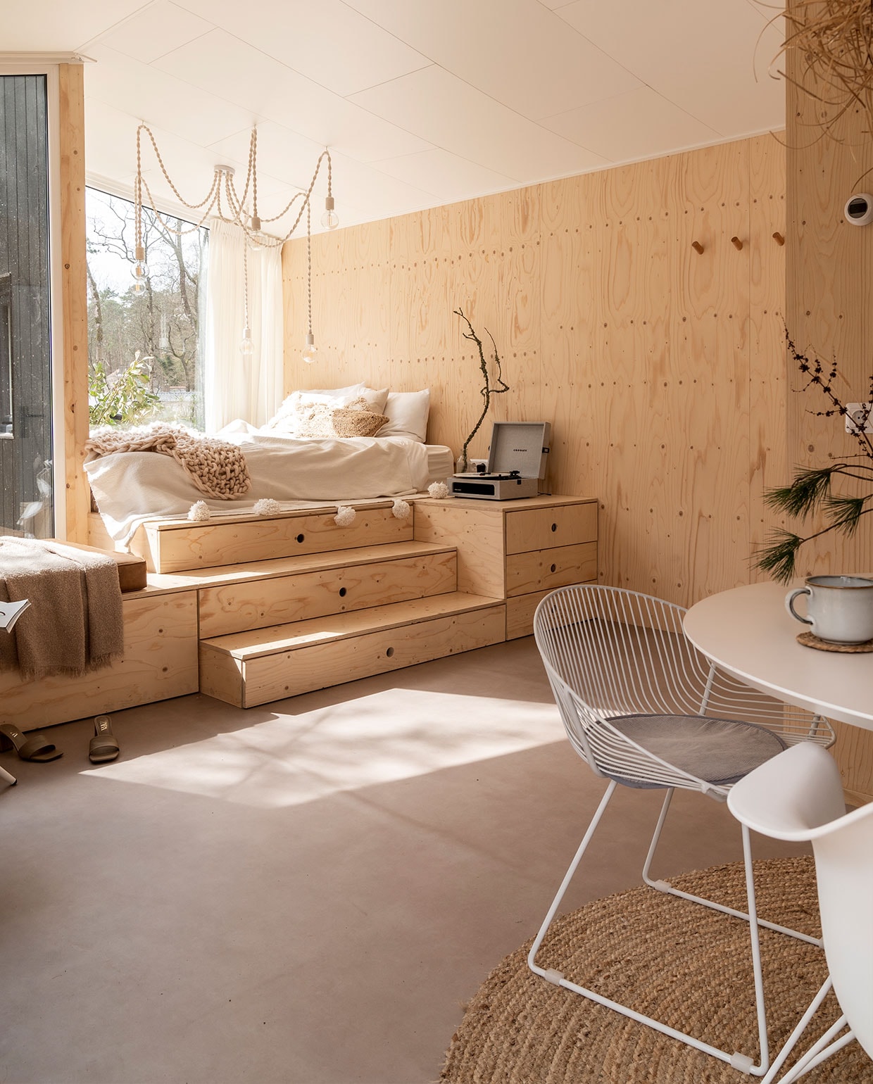 Bedombouw Hertog Recreatiebouw. Kandelaarlamp zelf­gemaakt. Matras Emma, beddengoed herkomst onbekend, sprei met pompons gekocht in Marrakech, gehaakte sprei Lidl. Waterkaraf Ikea, tak uit het bos, platenspeler Crosley, beige kussen H&amp;M Home, bruin kussen Calma House. Tafel Wehkamp, draadstoel, stoelen Vintagelab15. Mokken Ikea, droogbloemen Bloomon en uit het bos, kleed Volero Vloerkleden. vtwonen 11-2021 | Fotografie Muk van Lil | tekst Cathelijne van Wingerden