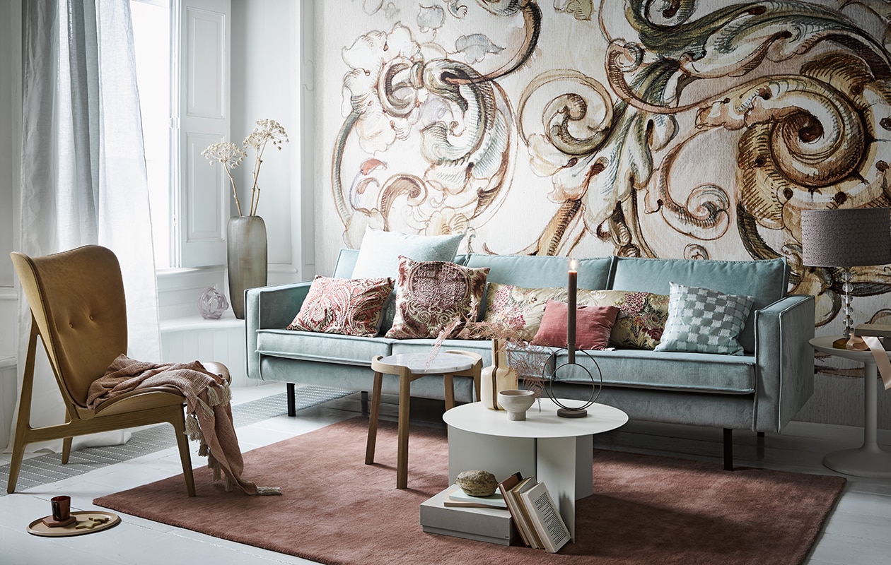 Behang Baroque R1151 (rebelwalls.com). Zeekleurige fluwelen driezitter Rodeo van BePureHome, 277 cm (vtwonen.nl). Deense fauteuil links, wit met gouden vaas op tafel, lampvoet rechts naast de bank, kaars, ronde kandelaar op tafel, mok op de grond, plaid op de fauteuil (loods5.nl). Witte stof Ditte voor het raam (ikea.nl). Kussens van links naar rechts alle stoffen per meter: Manipur van Designers Guild groot kussen van Designers Guild, kussen met bedrukte stof van Nobilis, langwerpig kussen van Nobilis-stof, blauw kussen, stof van Osborne &amp; Little, klein roze kussen Rivoli van Designers Guild (wva.nl). Lampenkap van stof Brooks van Zoffany, (barbaraemmy.com). Tafel met marmeren blad (bolia.com). Lage witte salontafel, roze kleed 140×200 cm, roze en blauw notitieboek, voorraaddoos op de grond, sierobject in raamkozijn (hay-amsterdam.com). Kom op salontafel (zarahome.com). Matglazen vaas met takken (homestede.nl). Overig privébezit. Beeld: vtwonen 03-2020 | styling Liza Wassenaar | fotografie Alexander van Berge