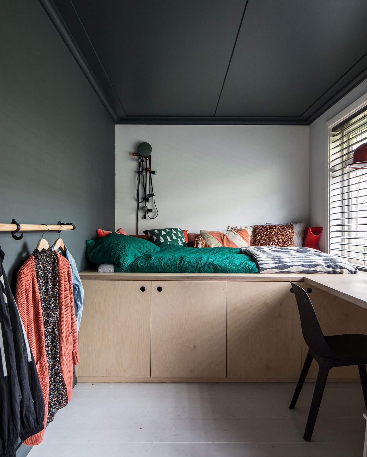Bed, kasten en bureau zelf ontworpen en gemaakt van multiplex. Kledingrail van oude steigerdelen. Op muur en plafond Full Marble, op de achterwand wit RAL 9016 en op de vloer trappenlak van Flexa. Vlinderjaloezieën Bece raamdecoratie, Hubo. Stoel Odger Ikea. Groene knop Dots Muuto, Flinders. Lichtstreng Kwantum. Dekbed-overtrek Hema. Sprei Ikea. Kussen rood, beige, bruin-roze en pluchen beagle Ikea. Groen kussen Ferm Living. Rood kussen La Redoute. Oranje-beige kussen HKliving. Rode lamp kringloop, design hanglamp Jieldé brocante markt Lille. vtwonen 10-2021 | Productie en Fotografie Corine Hoge Bavel | Tekst Marion de Boer