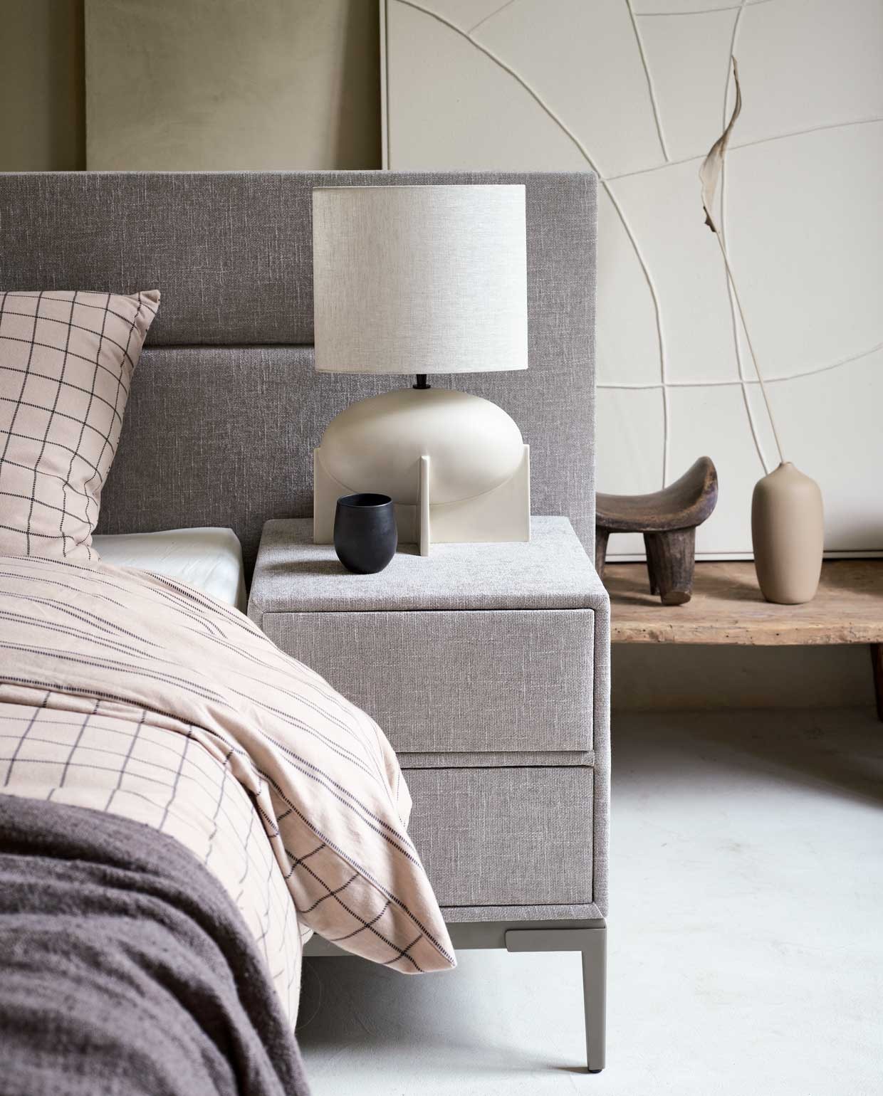 Boxspring Lifestyle by vtwonen Cinnamon Large vanaf € 1750,- (140×200 cm), nachtkastje Cardemom met twee lades, 45x45x62 cm (lxbxh) vanaf € 199,-, verkrijgbaar met verschillende stoffen en kleuren. Hier uitgevoerd met stof Mirage Taupe, metalen frame in kleur Champagne en poot Track. M.m.v. allestedlund.com (lamp), nordal.eu (grijze linnen plaid, kan en beker, marmeren schaal, linnen kussen), vtwonen.nl (zandkleurige vaas, beddengoed Grid in kleur zand uit de vtwonen collectie). vtwonen 10-2021 | styling Cleo Scheulderman | fotografie Jeroen van der Spek