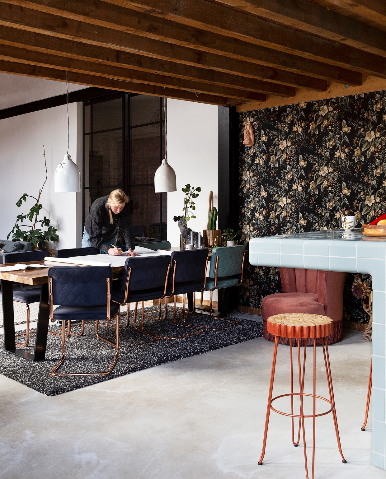 Martina aan de eetkamertafel in haar omgebouwde school in Wijk aan Zee. Eetkamertafel en stoelen Zuiver. Roze Dutchbone fauteuil Loods 5. Bufferlampen Pols Potten. Barkruk Kave Home Farley. Vloerkleed Ikea, behang Hornbach. Vloer van gevlinderd beton. vtwonen 10-2021 | styling Emmy van Dantzig | fotografie Jansje Klazinga
