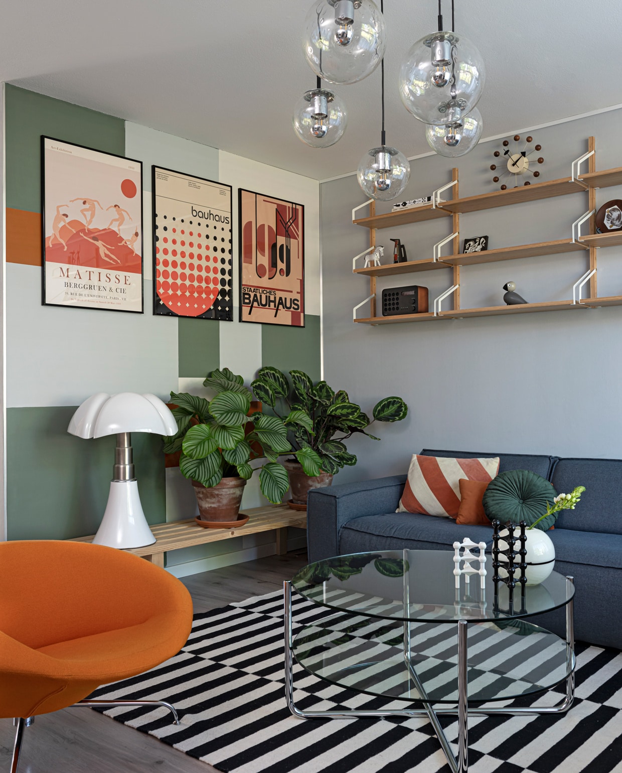 Hanglampen Raak Amsterdam via Marktplaats. Posters pstr.studio, lijsten Yourdecoration. Muren leemgroen en zinkgrijs Karwei, Indian spice Flexa. Plankjes Svalnäs Ikea. Met daarop: radio Eames, houten figuren Kay Bojesen (via Flinders), Ravelli keramiek rommelmarkt. Plantenpotten Intratuin, houten bankje zelf-gemaakt, tafellamp Pipistrello van Martinelli Luce via Marktplaats. Stoel Kirk van Artifort, glazen tafel Gispen via Marktplaats. Vloerkleed en vloer Ikea. Kandelaren Nagel via Catawiki, wit vaasje rommelmarkt. Bank Brooklyn Woon online, kussens Fonq, Loods 5 en Sissy-Boy Homeland. vtwonen 09-2021 | styling Sharon Roest | fotografie Caroline Martinot | tekst Sara Madou