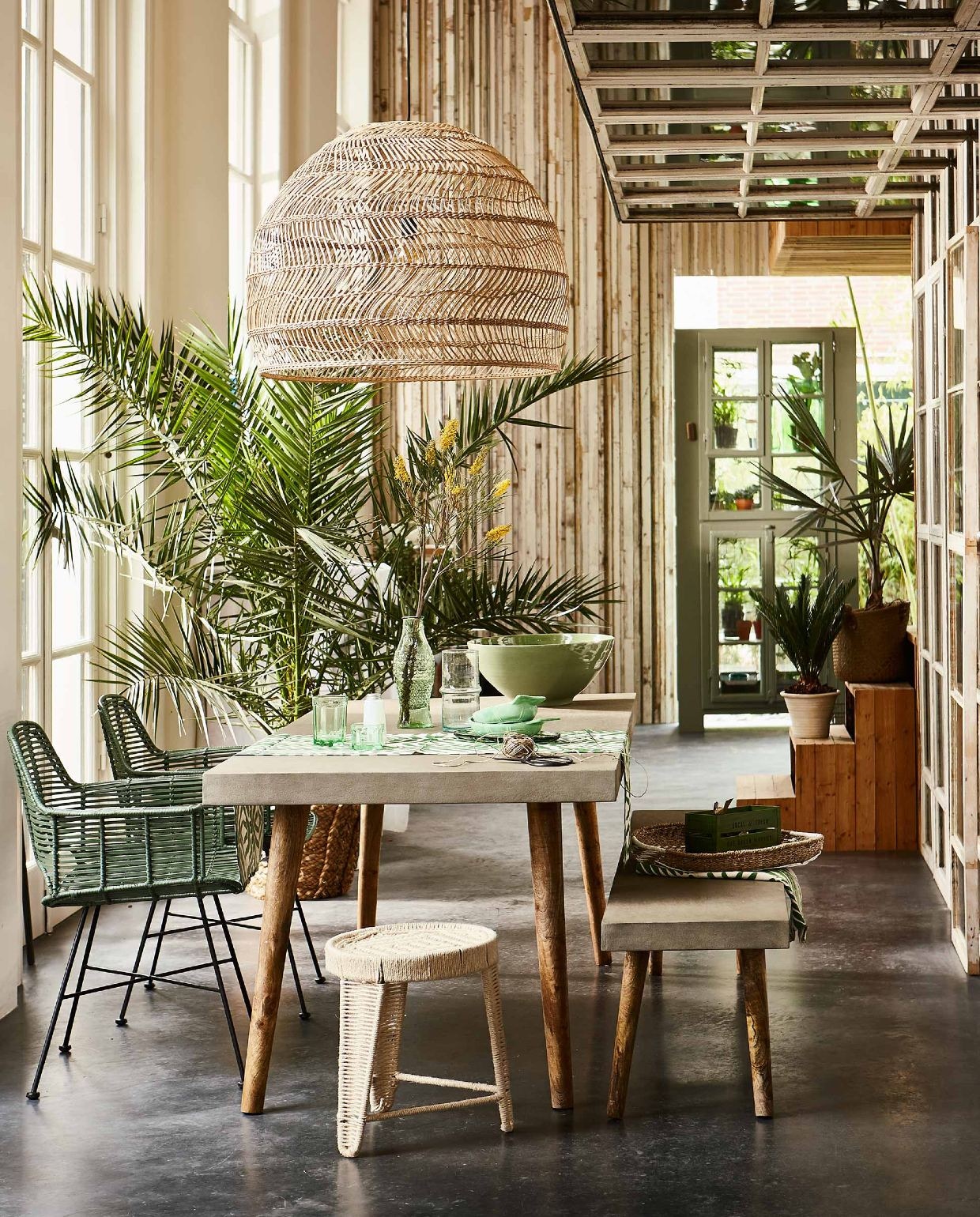 Tafel met betonnen blad en bijpassende bank (Nordal). Gevlochten hanglamp, groene rieten stoel (HK Living). Kruk van gevlochten touw, gevlochten schaal (Madam Stoltz). Lopers, groen met wit en groen kratje (H&amp;M Home). Karaf in de vorm van een vis, groene visvormige glazen schalen, lage visschaal van glas, laag groen glas, hoog groen glas, wit zoutvaatje, alles uit de collectie van Paola Navone (Serax). Glazen karaf met ribbel (Household Hardware). Grote groene aardewerken schaal (De Weldaad). Rieten mand met palm, gevlochten mand met palm op trap (Nordal). Groene kast (Combitex). Overig privébezit. vtwonen 07-2016 | Fotografie Jeroen van der Spek | Styling Cleo Scheulderman