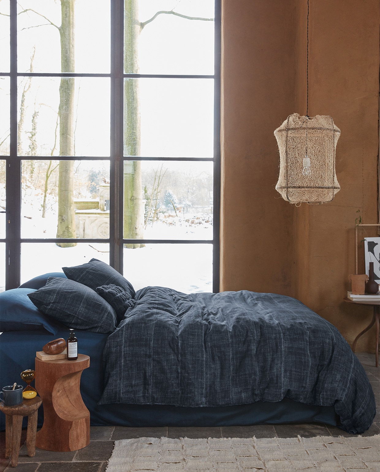 Kussen Tufted van katoen canvas, donkerblauw, 30×50 cm € 29,95. Dekbedovertrek Vintage Indigo van katoen, donkerblauw, 140×200/220 cm € 99,95, 200×200/220 cm € 189,95, 240×200/220 cm € 199,95.M.m.v. polspotten.nl (houten bijzettafel, donkerbruine vaas), studio-yen.com (vloerkleed, oude houten bijzettafel, houten steenvorm, keramieken pot), kasstoor.nl (ronde tafel driepoot), letoile.store (mok, lepel, zandloper, hanglamp, poster in lijst).Overig privébezit. vtwonen 09-2021 | productie Tanja Saarberg | styling Danielle Verheul | fotografie Richard Smit