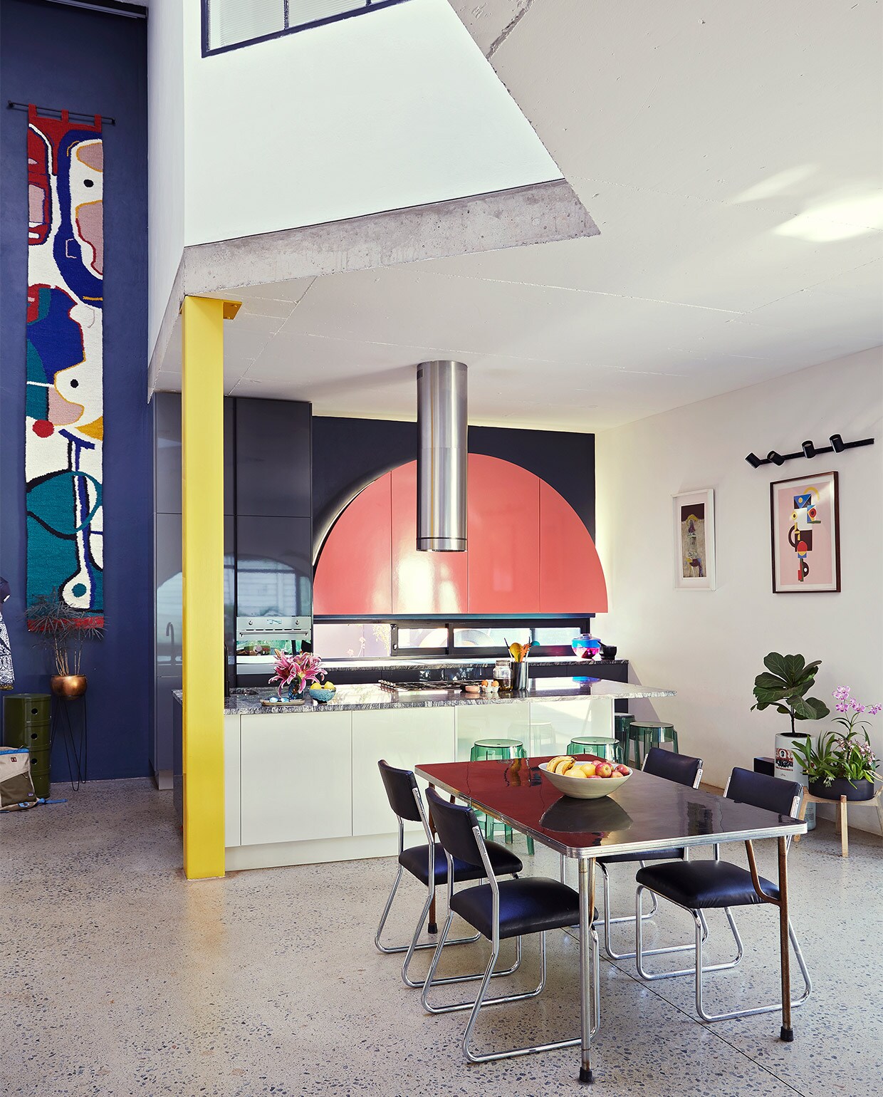Keuken ontworpen door Gregory Katz via ProReno Kitchens, met werkblad van Namibisch graniet International Slate Sales. Vloer gepolijst beton World of Decorative Concrete. Groene bijzettafel Componibili, barkrukken Charles Ghost en blauw-roze vaas Shibuya Kartell. Eettafel en chromen buisstoelen vintage. Fruitschaal Wonki Ware. Zwarte haken Leroy Merlin. Wandkleed Renée Rossouw. vtwonen binnenkijk special 07-2021 | productie Livinginside/Bureaux | fotografie\nElsa Young