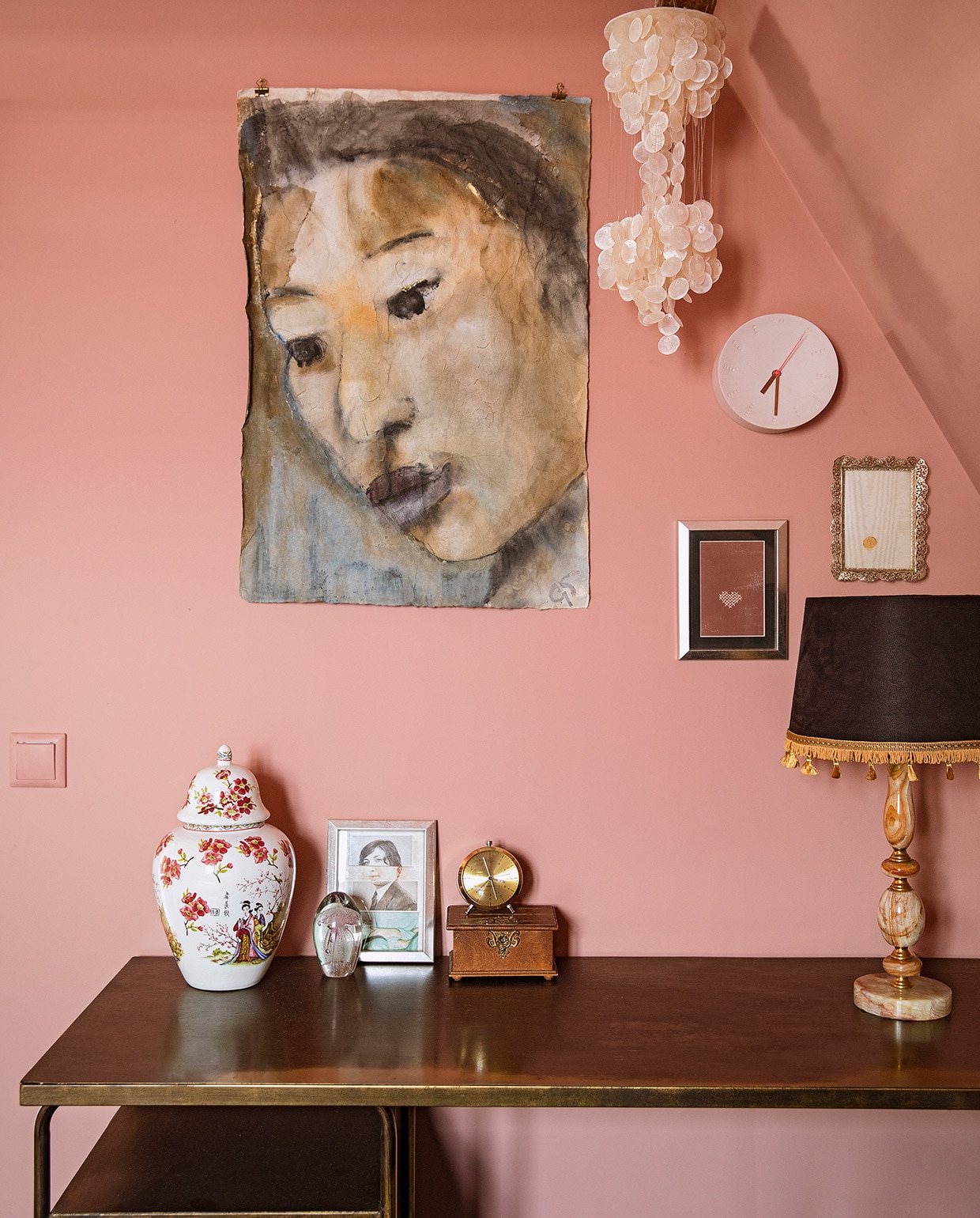 Muurverf Rosewood van Painting The Past. Sidetable BePureHome. Aardewerken pot, kwal in glas, gouden wekker en hangend schelpenornament herkomst onbekend. Ingelijste foto uitgeknipt uit tijdschrift Flow. Kistje en fotolijstjes erfstukken. Portret op doek Grace Spiegel. Wandklok Studio Harm en Elke. Zelfgemaakte tafellamp van tweedehands voet en gecustomized kap. vtwonen 06-2021 | Styling Ilona Jongepier | Fotografie Dana van Leeuwen | Tekst Renate Dahlmans