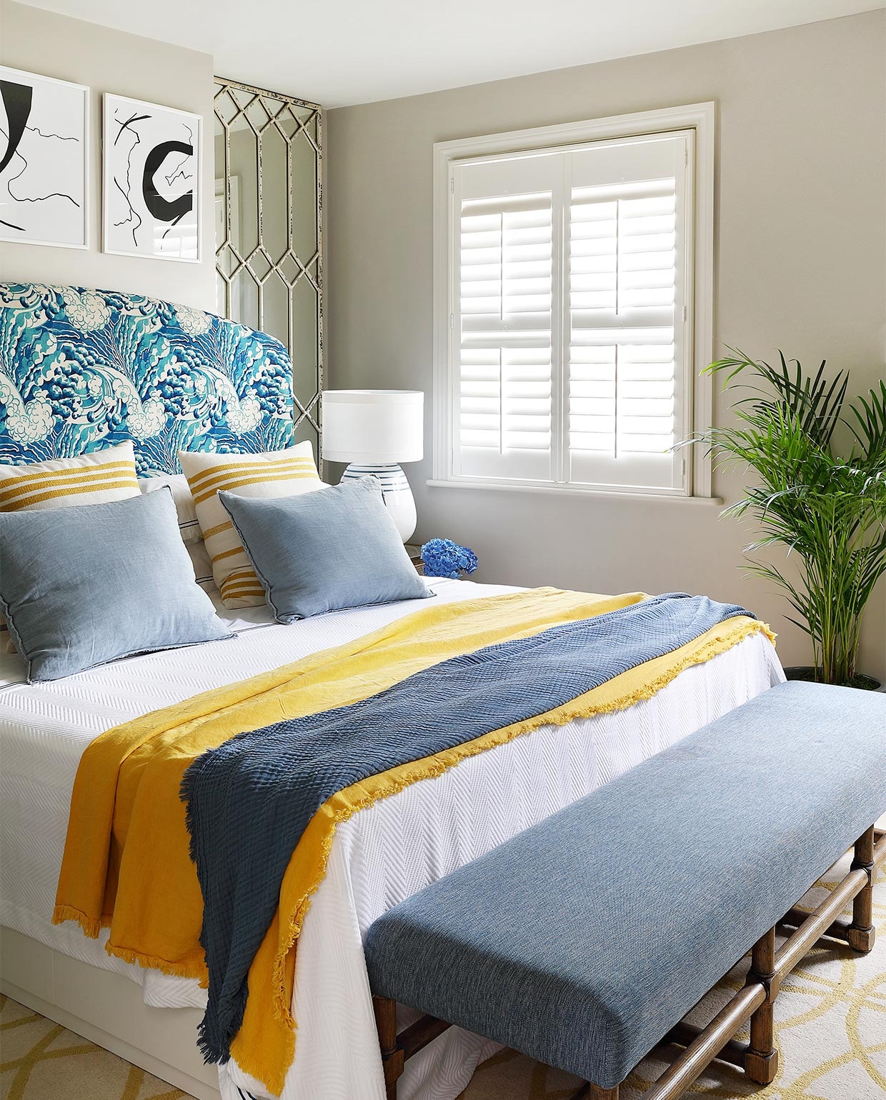 Muurverf Cornforth White van Farrow &amp; Ball. Het hoofdbord is bekleed met stof The Wave in de kleur mineral van Linwood Fabric. Beddengoed Granny Goose. Kussens met gele streep van stof Marina in kleur pastis van Lelièvre. Vaalblauwe linnen kussens lokaal gekocht. Gele en blauwe plaids H&amp;M Home. Spiegel Block &amp; Chisel. Tafellamp Gioalo van OKA. Bankje bekleed met stof van Mavromac. Kunst in lijsten gekocht via Etsy. vtwonen special zomerhuizen 07-2021 | productie Sven Alberding/Bureaux | fotografie Elsa Young/Bureaux