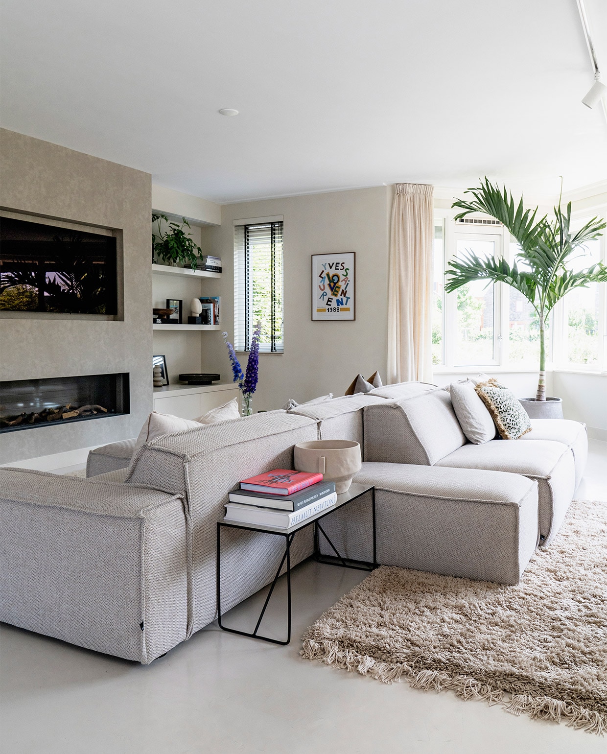 Vloer Tav Microcement. Modulaire bank Edge van Fest Amsterdam. Bijzettafel Broste Copenhagen. Vaas Zara Home. Televisie LG. Haard Dru. Poster aan de wand vintage. Kussens onder andere Nox en Loft. Plant Veitchia merrillii. Vloerkleed Westwing. vtwonen 08-2021 | fotografie Muk van Lil