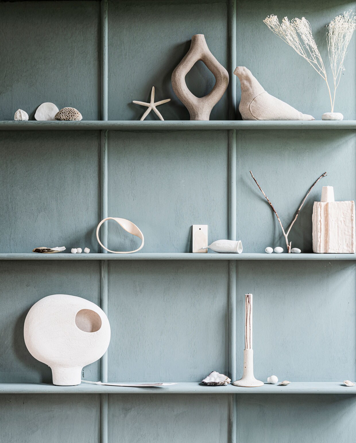 Kalkverf Blue Reef op de wand, per liter € 47,- (pure-original.nl). Grijs organisch gevormd kunstobject van Abid Javed € 389,-, cirkelvormig kunstobject van Slorence € 121,- (theodeto.com). Stoffen vogel € 45,- (homestede.nl). Keramieken vaas op middelste plank € 36,- (serax.com). Witte ronde vaas op poot van Urban Nature Culture, 26x7x27 cm (bxdxh) € 35,99 (sissy-boy.nl). Keramieken kandelaar € 34,90 (letoile.store). Porseleinen lampion € 44,-, porseleinen liaan € 45,-, porseleinen schelp € 35,-, porseleinen wandkelk € 70,-, porseleinen abutilon biscuit, zeewierparels, eucalyptus en drijfzaden, prijs op aanvraag (annekebruin.nl).Overig privébezit.