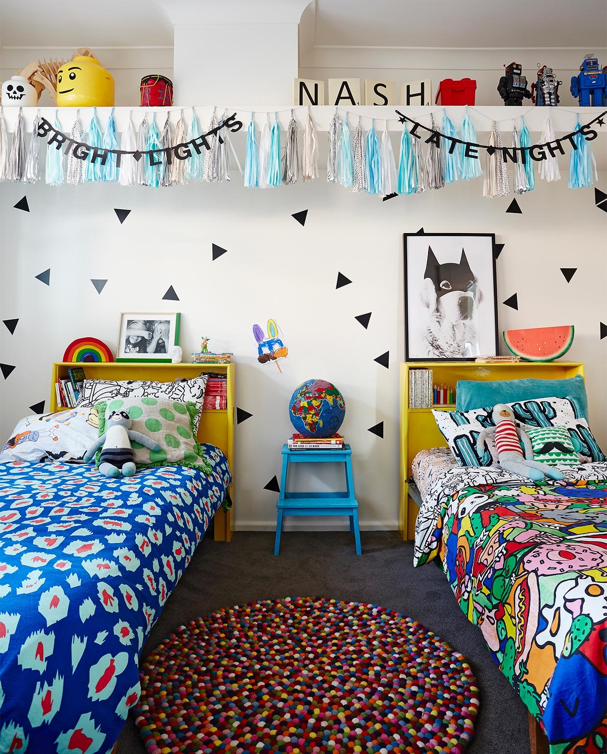 Muurstickers Urban Wall. Gele bedden gekregen van Hayleys tante, die er als kind in sliep. Dekbedovertrek rechts en zwart-wit kussen Kip &amp; Co. Trapje Bekväm Ikea, zelf blauw geverfd. Wereldbolkussen Hugg-a-Globe. Print Batdog My Dear Art Shop. Kwastenslinger DD Brand. Wit en geel opberghoofd Lego. Overig onbekend. vtwonen binnenkijken special 07-2021 | Styling Julia Green en Noel Coughlan | Fotografie Armelle Habib | Tekst Julia Green | Tekstbewerking Caroline Westdijk