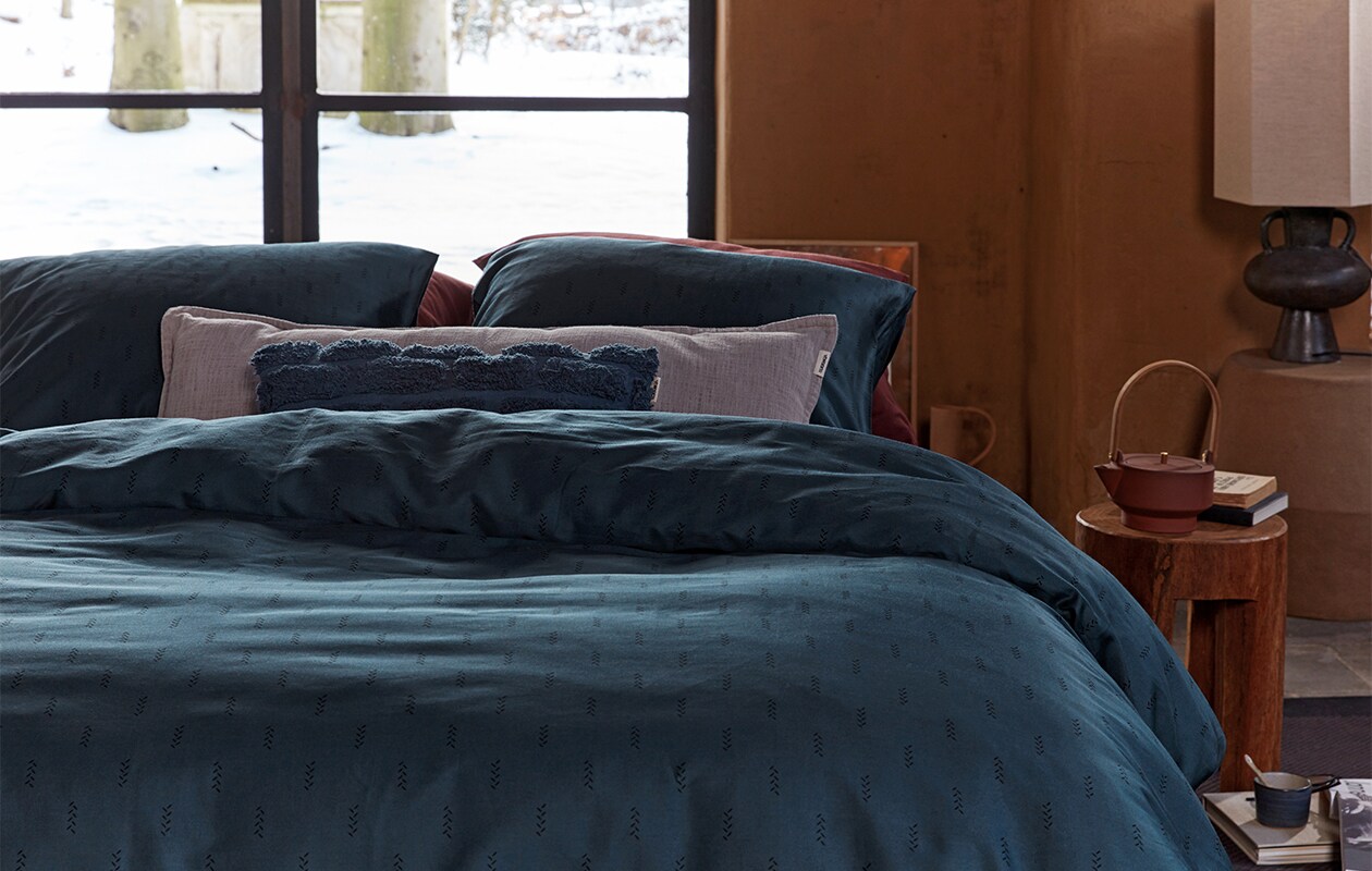 Kussen Tufted van katoen canvas, donkerblauw, 30×50 cm € 29,95. Dekbedovertrek Crops van katoensatijn, donkerblauw, 140×200/220 cm € 79,95, 200×200/220 cm € 149,95, 240×200/220 cm € 159,95 (ook in donkerrood).M.m.v. polspotten.nl (houten kruk), letoile.store (theepot, mok, lepel), loods5.nl/design (papier-maché bijzettafel in aardetint, tafellamp). Overig privébezit. vtwonen 09-2021 | productie Tanja Saarberg | styling Danielle Verheul | fotografie Richard Smit