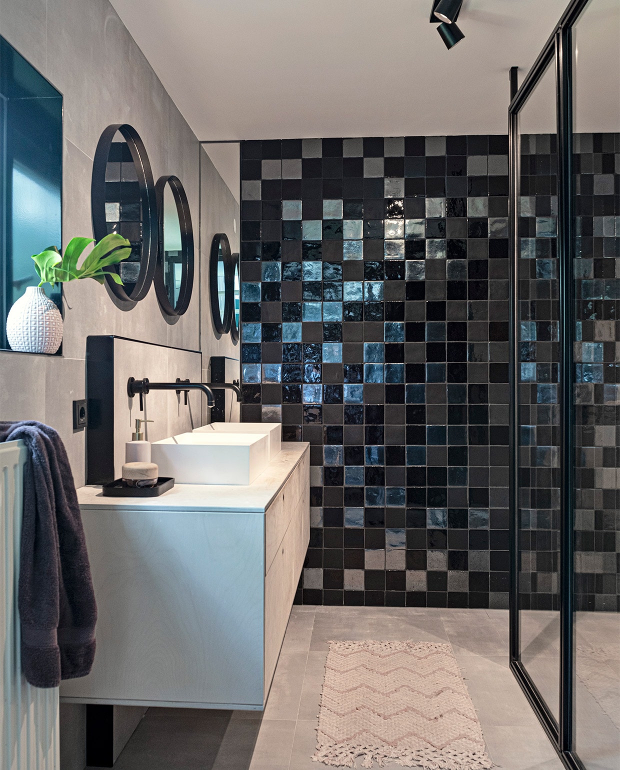 Grijze tegels op vloer en wand Spotlight van Villeroy &amp; Boch. Tegels achterwand Malaga in zwart-mat, zwart-glans en metallic Cottoceramix. Douchewand op maat gemaakt. Plafondspot Epic van Maretti. Composiet wasbak Dogma van Bliss Elements. Wandkranen uit de T-mix serie van Bongio. Wastafel van berken-multiplex door Leurs Interieurbouw. Spiegels Kwantum. Tray Zara Home. Badmat, handdoek, vaas en zeeppompje herkomst onbekend vtwonen 07-2021 | styling Sharon Roest | fotografie Caroline Martinot