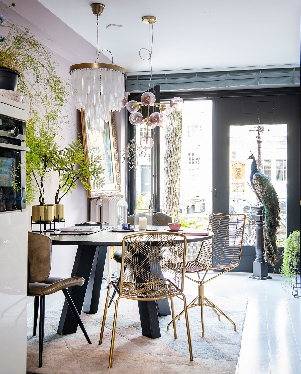 Ronde tafel van Leeelijk, leren stoelen van Wonen met Lef, de messing stoelen zijn van Zenza. Plantenhouders met messing potten Salix van Made, erin zijdeplanten van Silk-ka. Hanglamp met bollen van Made, lamp met hangende glazen pegels van Zenza. Vloerkleed van Rozenkelim, opgezette pauw van Taxidermy Amsterdam. Fles op tafel van Regalo Gallery, glas en roze kop-en-schotel van Anthropologie, blauwe waxinelichthouder van Zara Home. Het schilderij op de radiator is een souvenir uit Bali. vtwonen 06-2021 | productie Insidehomepage | styling Inge van Lieshout | fotografie Louis Lemaire