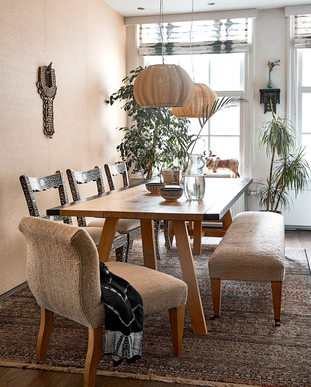 Keukentafel Heerenhuis Manufactuur, zelf verbreed. Stoelen uit India. Herkomst fauteuil en bank onbekend, bekleed met een hennepkleed uit Turkije. Op de fauteuil ligt een blouse. Zeegras behang uit Natural Wallcoverings collectie van Eijffinger. Schelpen- ornament, lampen en karaf Zenza. Wandkast uit Marokko, turquoise parfumflesvaas uit Turkije. Beeld koe souvenir uit India. Vloerkleed Dutchbone. Kommen Shabby Chic. Tie-dye raambekleding van een winkel die niet meer bestaat. vtwonen 06-2021 | productie Coco Features | styling Linda van der Ham | fotografie Peggy Janssen | Tekst Marlies Huisman, Mariska Keus