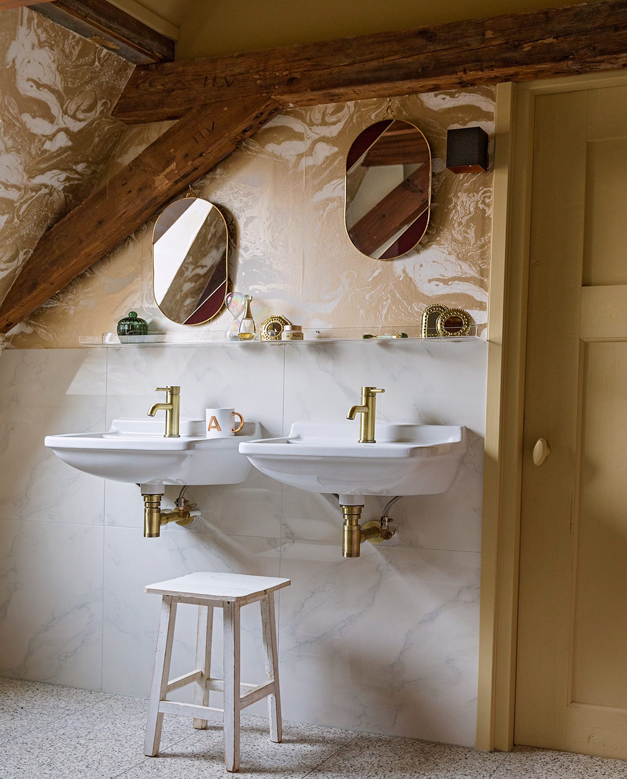 Terrazzovloertegels, keramische wandtegels met marmerprint, kraan van geborsteld koper en witte kruk herkomst onbekend. Behang Marble Gold 188 van Ferm Living. Verfkleur houtwerk Bisquit van Painting The Past. Wastafels Villeroy &amp; Boch. Plexiglas wandplankjes Ikea. Ovalen spiegels &amp;Klevering. Mok met gouden oor cadeau gekregen. vtwonen 06-2021 | styling Ilona Jongepier | fotografie Dana van Leeuwen