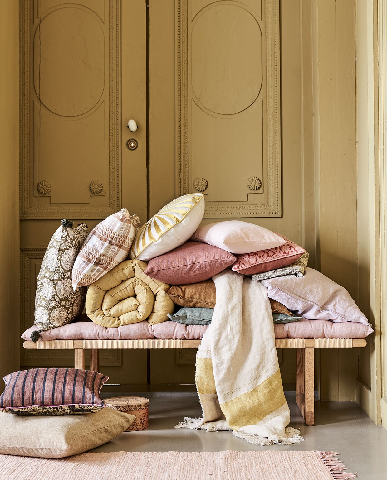Bankje BM0488 van Børge Mogensen &amp; Carl Hansen, 138x46x34,5 cm (bxdxh) € 1440,-, geruit kussen € 49,90, plaid met streep van Libeco,110×180 cm € 122,-, donkerroze kussen, 40×80 cm € 79,90, klein roze kussen, 30×40 cm € 44,90, (letoile.store). Roze vloerkleed Chindi Mat Rose, 60×90 cm € 20,-, kussen linksonder op de grond, 50×50 cm € 45,-, kussen met print links Shimla Olive, 50×50 cm € 47,-, geel matras Mirra Lemon Curd, 55×150 cm € 55,-, roze matras Mirra Rose, 55×150 cm € 55,-, rond kussen met geel borduurwerk € 95,-, groen kussen, 50×50 cm € 47,-, oker kussen, 50×50 cm € 47,-, lila kussen, 50×50 cm € 47,-, groen tafelkleed met print, 150×250 cm € 100,-, roze kussen met borduurwerk bloemen, 30×40 cm € 53,75, alles van Bungalow (scandicliving.nl). Kussen met strepen linksonder Doris van HKliving, 30×40 cm € 57,95 (hkliving.nl). Doosje € 3,- (jolandelijkdewebshop.nl). Kleur op de wand vergelijkbaar met Gervase Yellow No.72 van Farrow &amp; Ball. vtwonen 05-2021 | styling Marlies Does | fotografie Bart Brussee