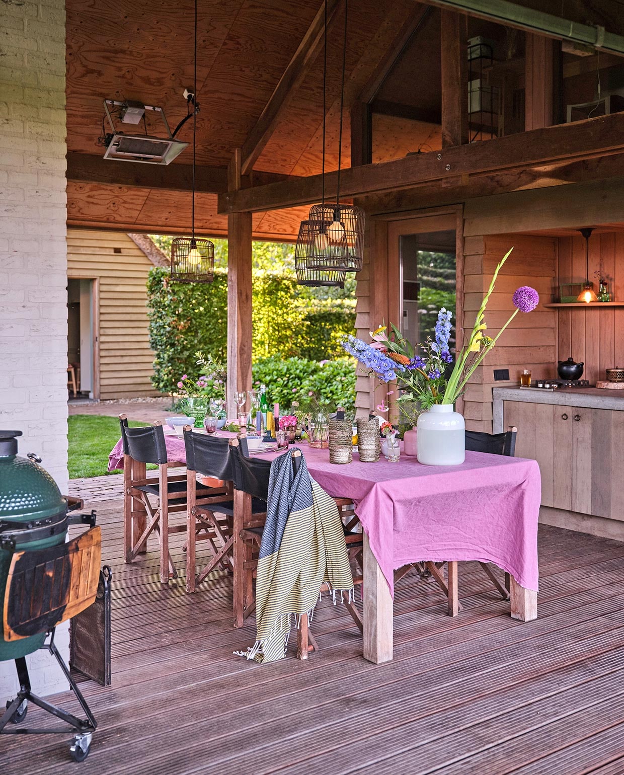 Keuken in tuinhuis zelfgemaakt van wrc-delen, rode ceder. De lampen zijn vogelkooien die zwart gespoten zijn. Het roze tafellaken is een onafgewerkte linnen lap van de markt. Barbecue Green Egg. Hammamdoek Sissy-Boy Homeland. Regisseursstoelen Vervoort Meubelen. vtwonen tuin 03-2021 | styling Carolien Schrievers | fotografie René van der Hulst