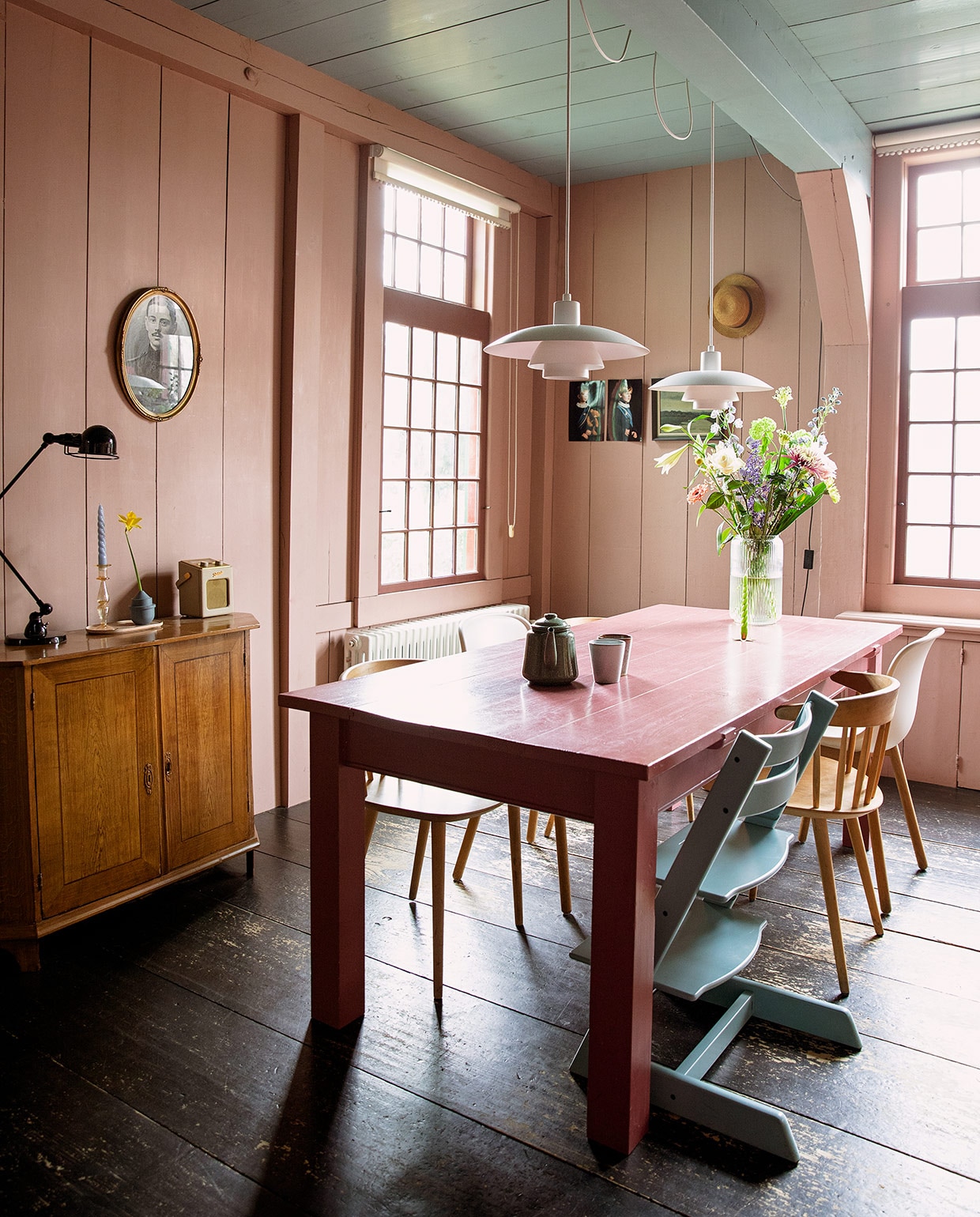 Muurverf lijnolieverf van Aquamarijn gemengd met Sulking Room Pink van Farrow &amp; Ball, kleurnummer lijnolieverf op plafond onbekend, beide via Rigo Verffabriek. Rolgordijn Hansa, kast antiek, bureaulamp Jieldé, kandelaar en groene kaars Anna + Nina. Blauwe kaars, tray, witte stoel en houten stoelen Hay. Kinderstoel Stokke. Blauw vaasje Mickey Philips, radio Roberts. Ovaal portret van overgrootvader Bart, portretten van Tom en Siewert gemaakt door een moeder van school, schilderij van Jan de Boer erfstuk, oude hoed van Bart. Hanglampen Louis Poulsen. Tafel vintage, vaas en kopjes Sissy-Boy, theepot Dille &amp; Kamille. vtwonen 05-2021 | productie en Styling Ilona Jongepier | fotografie Dana van Leeuwen