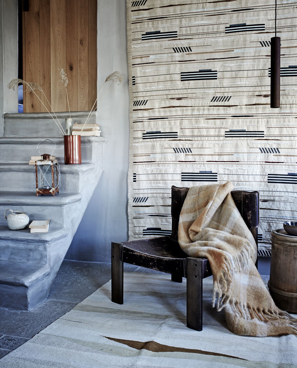 Stof wandkleed Pelops Gris Ombre van Nobilis, vloerkleed Vegetal Natural Stripes van Golran (wva.nl). Vintage leren fauteuil (pietjonker.nl). Mohair deken met ruit (zarahome.com). Kan, ecru vaas, schaaltje, alles van Serax (vtwonen.nl). Windlicht (com-anders.nl). Rode cilindervaas (klevering.nl). Houten bijzettafel (burbri.nl). Overig privébezit.