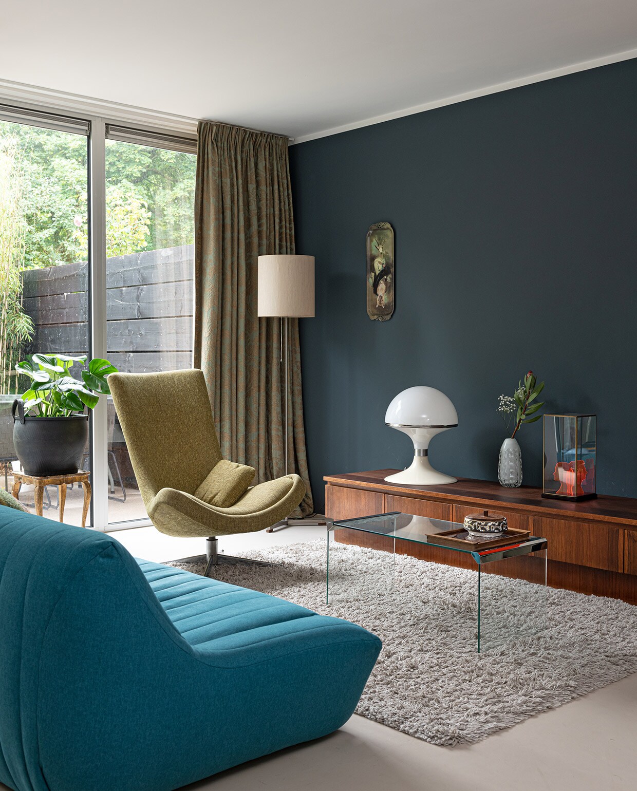 Muurverf Deep Ocean van Histor, gordijnen van de moeder van Charlotte. Staande lamp, tafel tweedehands. Plantenpot Household Hardware. Draaistoel Rudolf Wolf. Bank Made. Kleed Shaggy van Trendcarpet. Marmoleumvloer Walton Forbo vloeren. Dressoir vintagevirus.nl. Lamp Concept van Brabantia. Op het dressoir: middelste vaas kringloop-winkel. Object rechts eigen ontwerp. Portret in vitrine HKliving. Glazen tafeltje Marktplaats. Keramieken bakje, dienblad vintage. Plateau madeindesign.com . vtwonen 04-2021 | styling Sharon Roest | fotografie Caroline Martinot