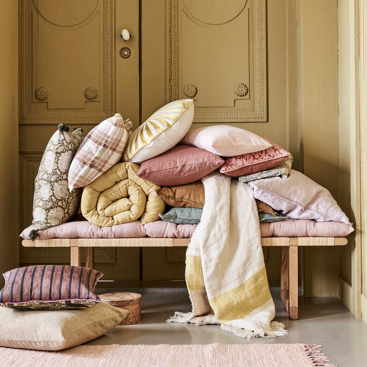 Bankje BM0488 van Børge Mogensen &amp; Carl Hansen, 138x46x34,5 cm (bxdxh) € 1440,-, geruit kussen € 49,90, plaid met streep van Libeco,110×180 cm € 122,-, donkerroze kussen, 40×80 cm € 79,90, klein roze kussen, 30×40 cm € 44,90, (letoile.store). Roze vloerkleed Chindi Mat Rose, 60×90 cm € 20,-, kussen linksonder op de grond, 50×50 cm € 45,-, kussen met print links Shimla Olive, 50×50 cm € 47,-, geel matras Mirra Lemon Curd, 55×150 cm € 55,-, roze matras Mirra Rose, 55×150 cm € 55,-, rond kussen met geel borduurwerk € 95,-, groen kussen, 50×50 cm € 47,-, oker kussen, 50×50 cm € 47,-, lila kussen, 50×50 cm € 47,-, groen tafelkleed met print, 150×250 cm € 100,-, roze kussen met borduurwerk bloemen, 30×40 cm € 53,75, alles van Bungalow (scandicliving.nl). Kussen met strepen linksonder Doris van HKliving, 30×40 cm € 57,95 (hkliving.nl). Doosje € 3,- (jolandelijkdewebshop.nl). Kleur op de wand vergelijkbaar met Gervase Yellow No.72 van Farrow &amp; Ball vtwonen 05-2021 | styling Marlies Does | fotografie Bart Brussee