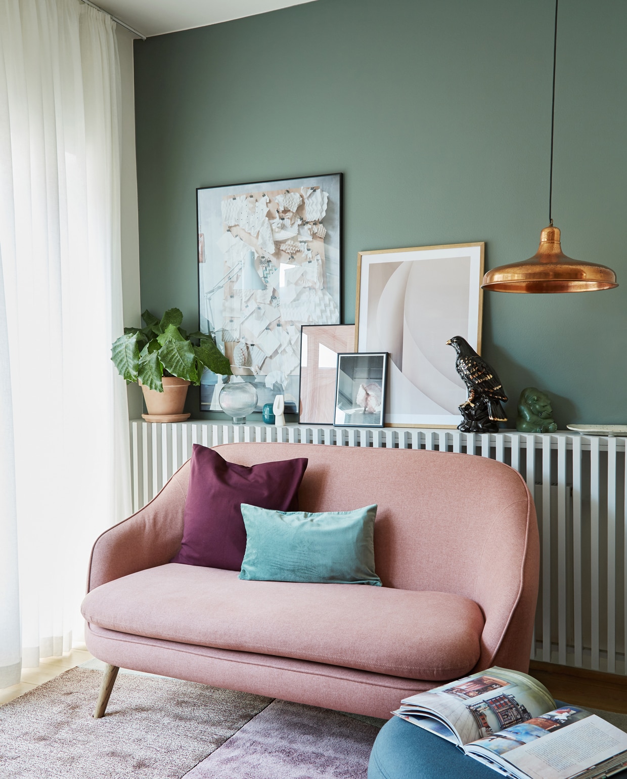 Vloerkleed van Please wait to be seated, gordijnstof Jab. Bank van Sofacompany. Radiatorombouw zelfgemaakt voor items die ze nergens kwijt konden in het huis zonder vensterbanken. Grote poster werk van Anitta uit het boek Arts and Crafts Homes, roze print en lichte poster By Garmi, klein plaatje Dos Family. Kussens H&amp;M Home. Lamp vlooienmarktvondst. Poef Warm Nordic. Muurverf Vineyard nr. 61 van Flügger. vtwonen 02-2020 | styling Rikke Graff Juel | fotografie Anitta Behrendt/Living Inside