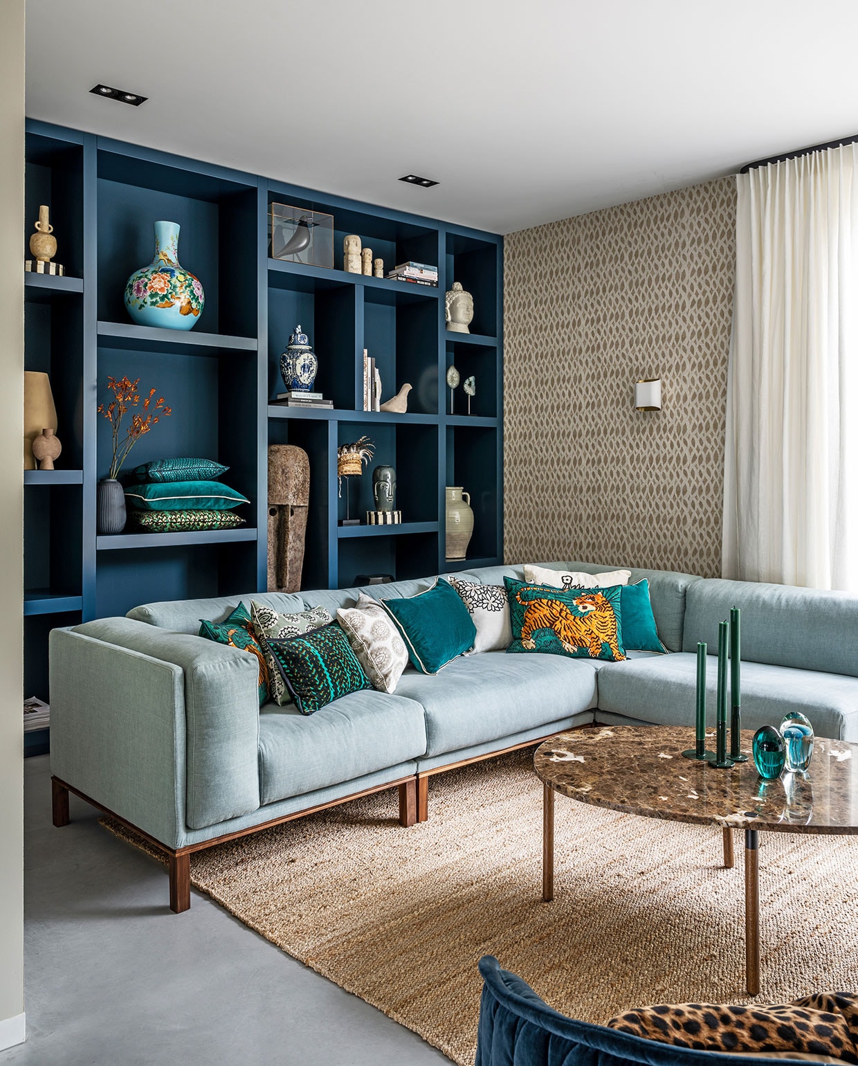 Vakkenkast Maek Meubels in kleur Hague Blue van Farrow & Ball, behang Perles Topaze van Élitis, linnen gordijnen van Interstil, bank en salontafel Bolia, vloerkleed TineK Home, kussens Élitis, kaarsen Sissy-Boy, geëmailleerde groene kaarsenstandaards Tica. In kast: lichtblauwe vaas Pols Potten, zandkleurige vaas en bruine vaas Casa Gitane, witte vogel Pigeon van Christien Meindertsma, zwarte vogel House Bird Vitra Eames, witte figuren rechtsboven Stone Man Dinor en hoed via Loods 5, overig privebezit.