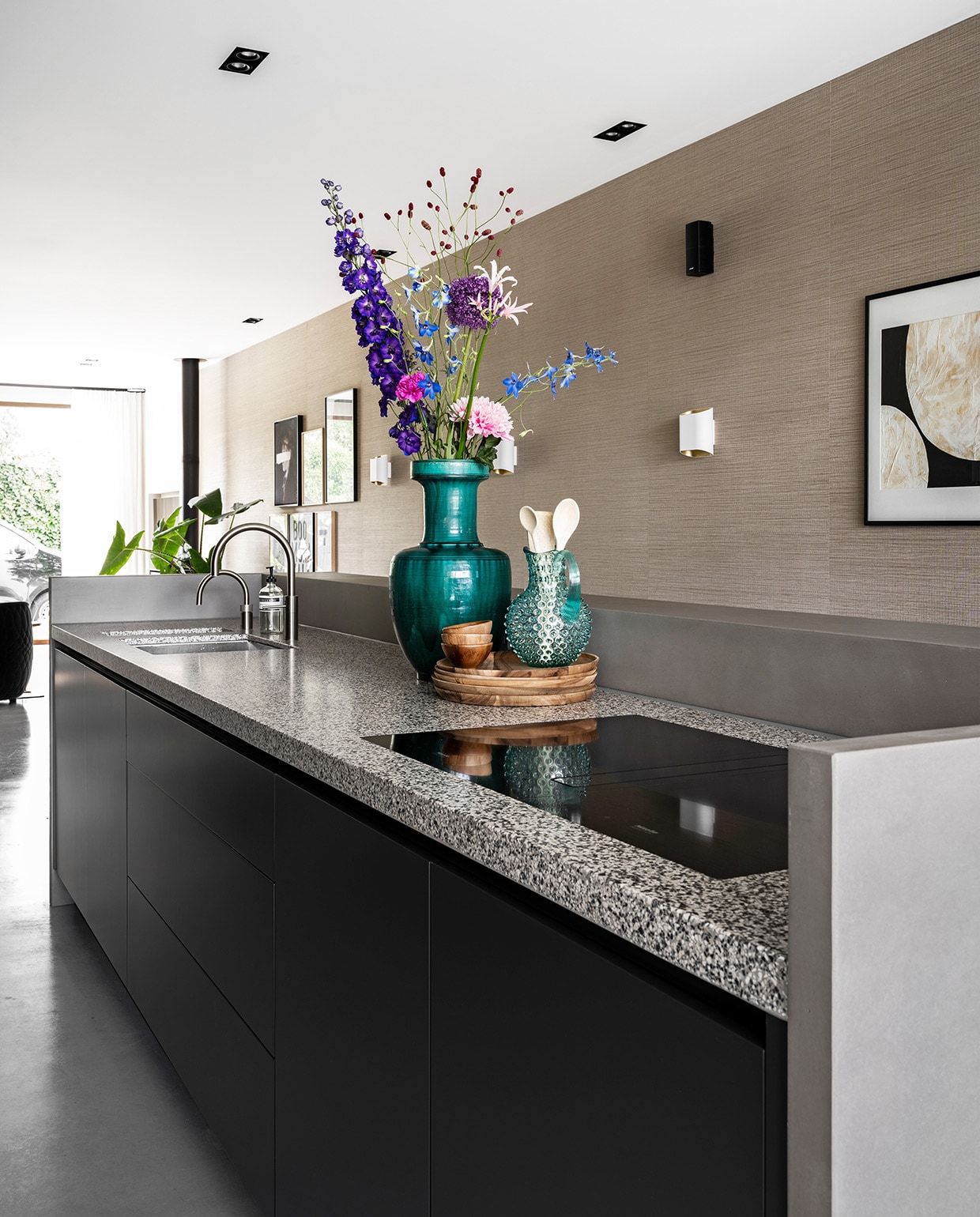 Zelf ontworpen keuken met granieten aanrechtblad en betonnen rand gemaakt door Maek Meubels, apparatuur Miele, blauwgroene vaas en blauwe kan Mischa Hulshof Alkmaar, houten borden en schalen vtwonen collectie, kunstwerk aan muur To the moon and back van Art in Return, witte wandlamp Trapz Modular Lighting Instruments. vtwonen 03-2021 | productie CocoFeatures | Styling Wendy Verhaegh | fotografie Stan Koolen