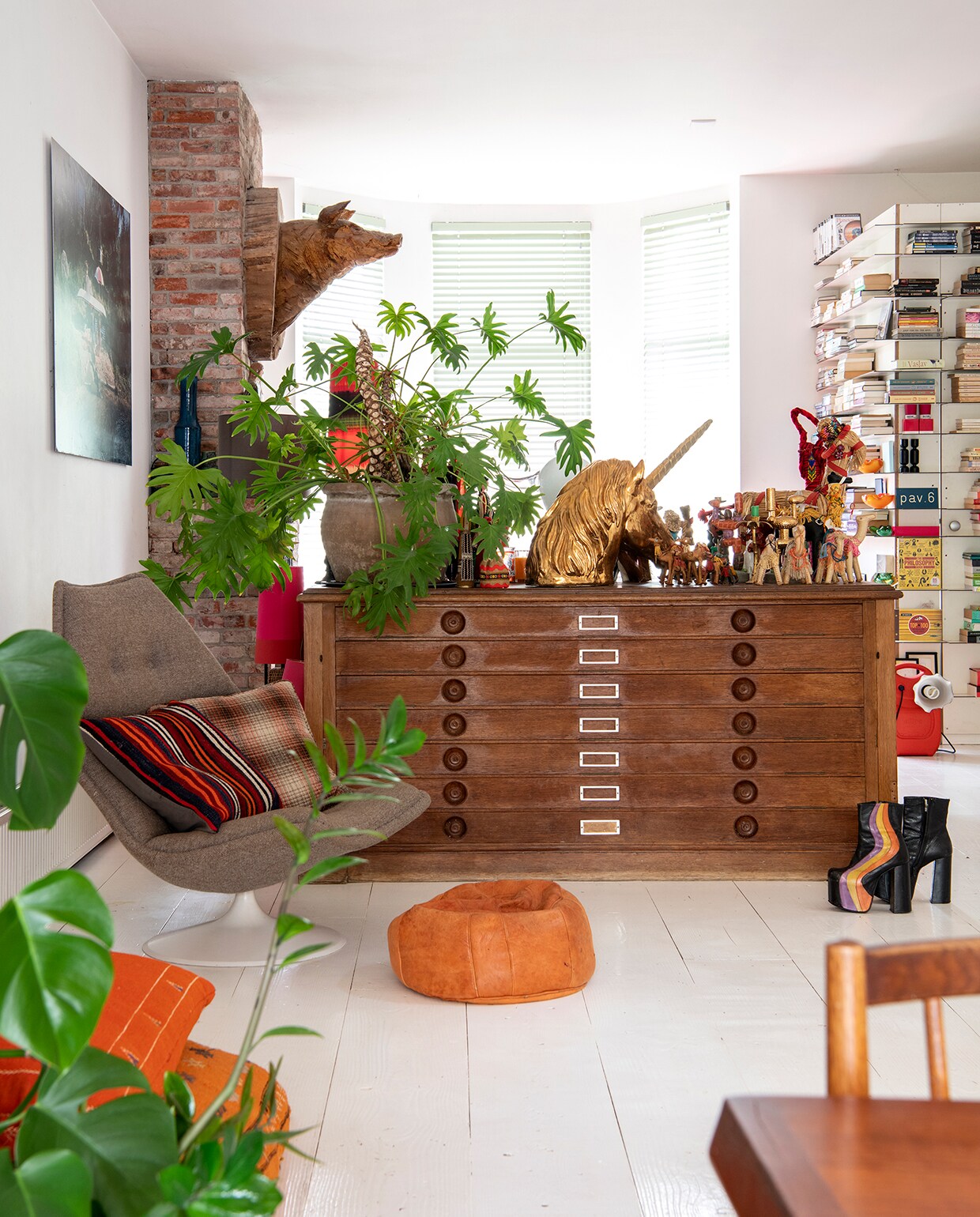 Ladekast gekocht van een kunstenaar. Plantenpot tuincentrum. Gouden eenhoorn Hans van Bentem. Collectie vintage (dieren)beelden. Vintage fauteuil Artifort. Kelimkussen Ikea, geruit kussen vintage, grote vloerkussens zelfgemaakt van Marokkaanse kelims. Leren poef Loods 5. Houten torso van varken gekocht op festival. Kunstwerk aan de muur gemaakt door Karel. Rode lamp Laura de Monchy. Boekenkast ontworpen door Karel.