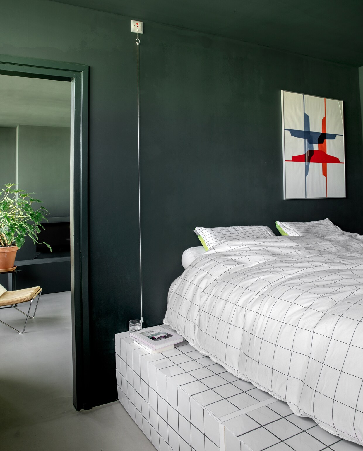 Doorkijkje naar de woonkamer vanuit de slaapkamer. Het plateau waarop het bed staat, werd gebouwd van mdf en vervolgens betegeld. Chantal: ‘Die donkere kleur op muren en plafond maakt het extra knus. Deze kamer voelt als een soort mega bedstee.’ Vintage Bachelor Chair van Verner Panton via Niet Nieuw Design. Plant en terracottapot Intratuin. Witte tegels 10×10 cm Hornbach. Boek Little Escapes via bartsboekje.com. vtwonen binnenkijkspecial 12-2019 | styling José Martens | fotografie Peggy Janssen