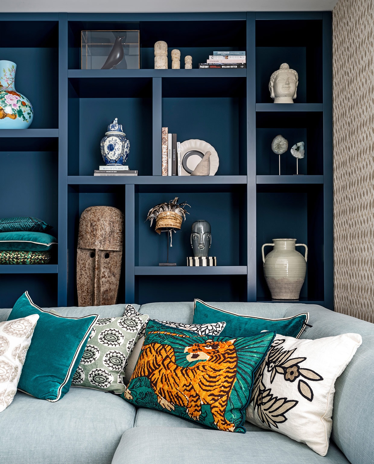 Vakkenkast gemaakt door Maek Meubels Heerhugowaard geschilderd in de kleur Hague Blue van Farrow &amp; Ball, behang Perles Topaze van Élitis bank Bolia, kussens Élitis. In kast: lichtblauwe vaas Pols Potten, zandkleurige vaas en kleine bruine vaas Casa Gitane, witte vogel Pigeon van Christien Meindertsma, zwarte vogel House Bird Vitra Eames, witte figuren rechtsboven Stone Man Dinor en hoed op standaard Bali via Loods 5, overige items souvenirs en cadeaus. vtwonen 03-2021 | productie CocoFeatures | styling Wendy Verhaegh | fotografie Stan Koolen
