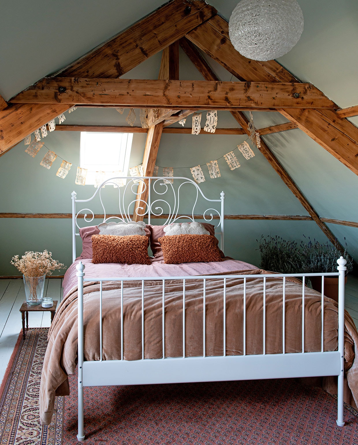 Muurverf Vert de Terre van Farrow &amp; Ball. Witte lak op vloer Action. Slinger zelfgemaakt van oude boeken. Hanglamp Kwantum. Bed Ikea. Lakens Jysk. Nachtkastje, vloerkleed en beige kussens kringloopwinkel. Vaas gekregen. Bloemen zelf gedroogd. Geurkaars Sissy-Boy. Terracotta kussens Jysk. Plaid H272984&amp;M Home. Lavendel uit de tuin van familie. Potten Tuincentrum Coppelmans. vtwonen 03-2021 | fotografie Caroline Coehorst