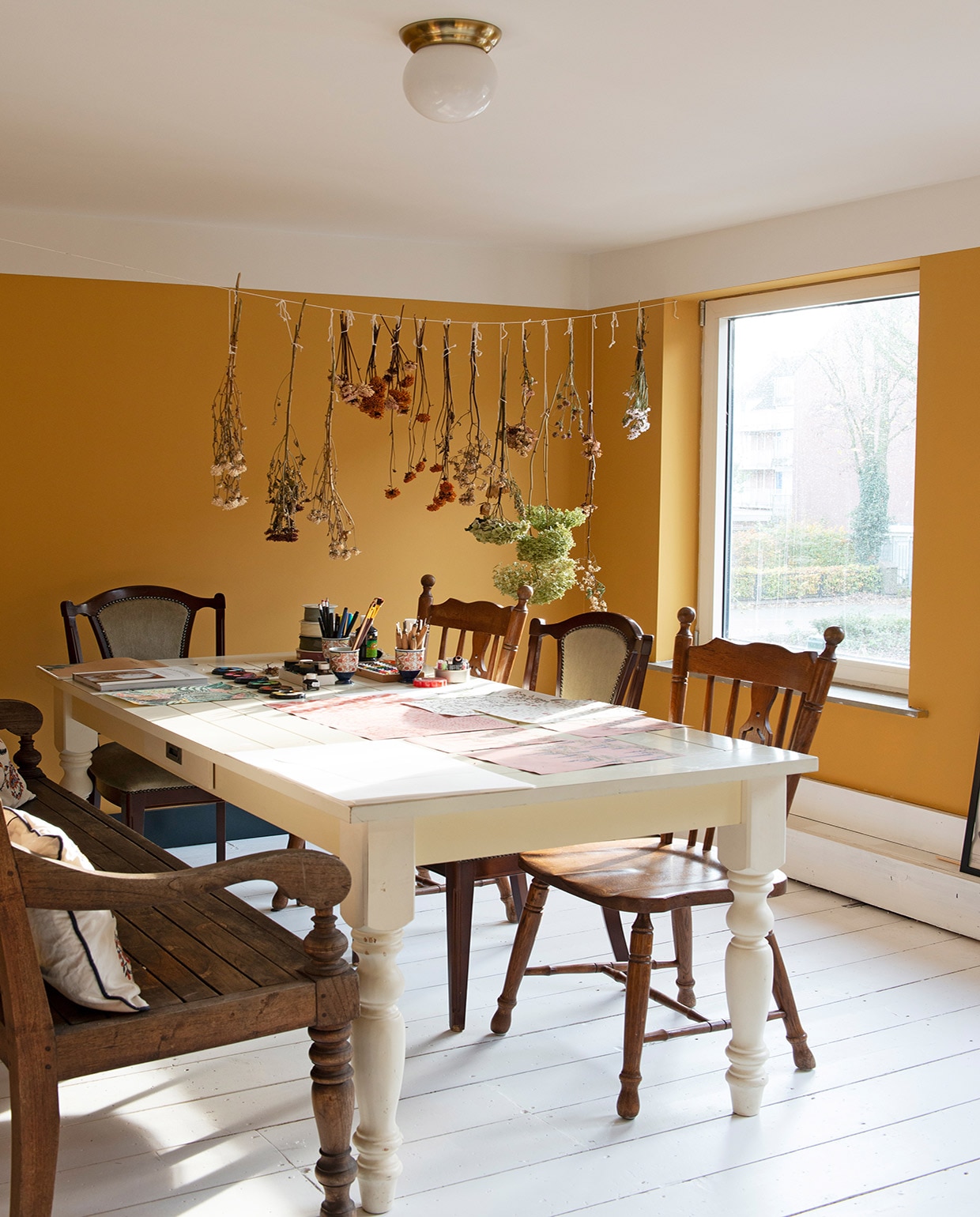 Muurverf India Yellow van Farrow &amp; Ball. Verf plafond Slaked Lime van Little Greene. Vloer origineel. Plafondlamp Ikea. Bloemen zelf gedroogd. Tuinbank gekregen, kussens Ikea. Tafel Marktplaats. Gestoffeerde stoelen kringloopwinkel. Houten stoelen en bekers gekregen. vtwonen 03-2021 | fotografie Caroline Coehorst