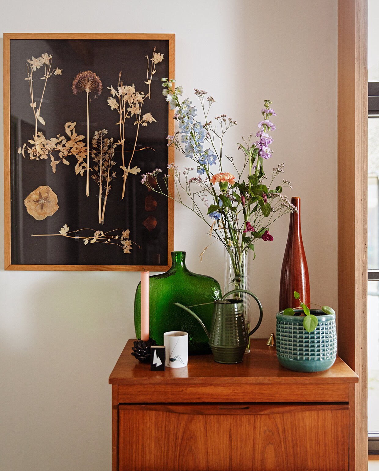 Kastje vintage. Droogbloemen in lijst gemaakt door Femke. Blauwe vaas tuincentrum. Rode vaas H&amp;M Home. Groene vaas rommelmarkt. Gieter Nordic Nest. Kandelaar Koekwaus. Witte kaarsenhouder Anna + Nina. vtwonen 03-2021 | Styling Ilona Jongepier | Fotografie Dana van Leeuwen | Tekst Sara Madou\n\nvtwonen 03-2021 | kast met vazen en kunst aan de muur