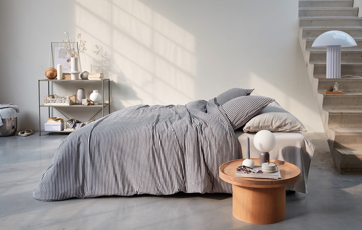 Katoen jersey dekbedovertrek Comfy Stripe in kleur grijs, 140×200/220 cm en 240×200/220 cm (inclusief kussenslopen). Ook verkrijgbaar in kleur donkergrijs, lichtgrijs en duotoon oker. M.m.v. vtwonen.nl (witte lamp van HKliving, houten bijzettafel, nachtlamp van Bolia), letoile.store (poster in lijst, grijs bolvaasje, witte kratjes, karaf met glas), loods5.nl (wandkast, witte vazen, grijze langwerpige vaas, witte bolkandelaar, keramieken opberger met deksel). Overig privébezit. vtwonen 04-2021 | productie Tanja Saarberg  | styling Danielle Verheul | fotografie Richard Smit
