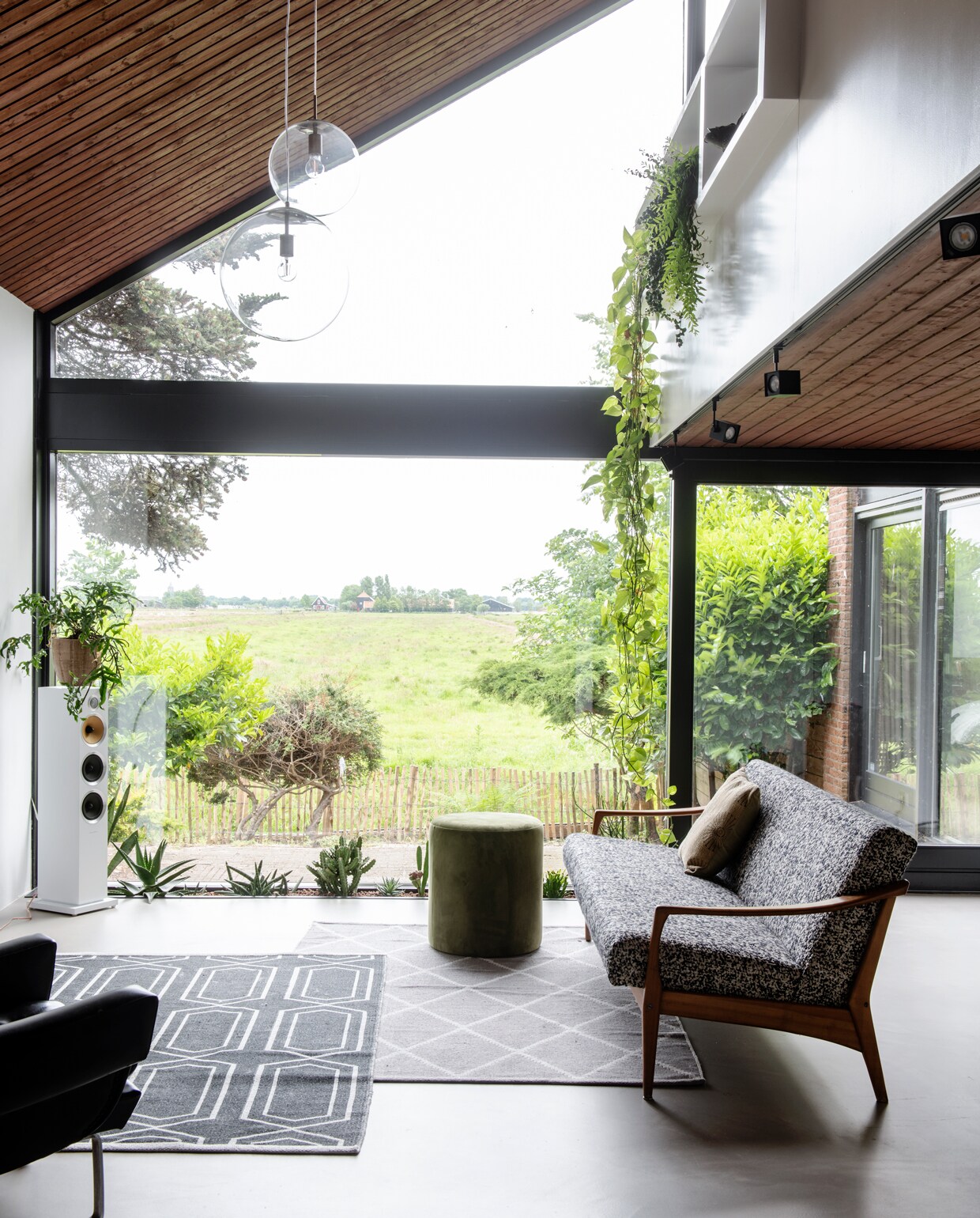 De woonkamer met uitzicht op de polder. Vloerkleden H&amp;M, vintage bank Decennia Design, groene poef van Riverdale, witte speaker Bowers &amp; Wilkins, bollampen van Rietveld Licht &amp; Wonen, spots Altra Dice via slv.com. De oorspronkelijke convectorput doet nu dienst als XL plantenbak. vtwonen 01-2020 | styling Kim Timmerman/Insidehomepage | fotografie Louis Lemaire