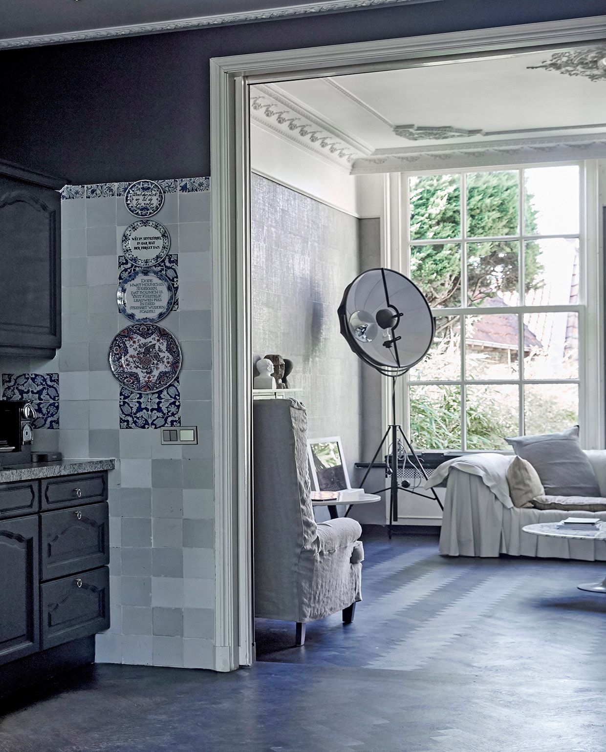 Keuken donkerblauw geschilderd. Tegels op achterwand originele Harlinger tegels. Borden met Friese spreuken aan muur vintage. Doorkijkje naar de achterkamer: stalamp Fortuny van Pallucco, leren bank Bohémien van Busnelli, kussens op bank gemaakt van stoffen van Emery &amp; Cie. Witte tv Bang &amp; Olufsen. Witte buste van een winkel die niet meer bestaat, antieke houten buste uit India, zwart hoofd van GMC uit Zuid-Afrika. Lage marmeren tafel Knoll, hoge marmeren tafel Tom Dixon. vtwonen 02-2021 | Styling Marianne Luning | Fotografie Tjitske van Leeuwen | Tekst Simone Braaksma