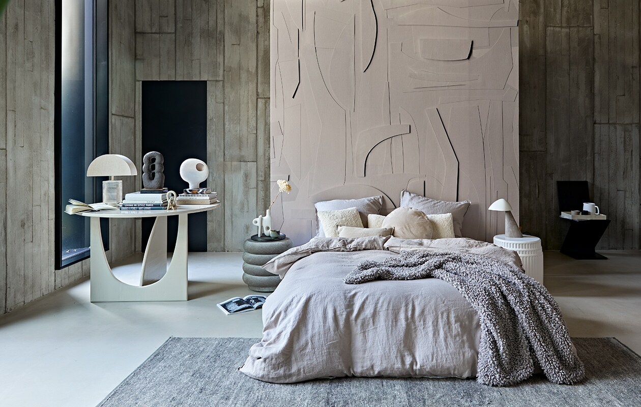 Behang op roomdivider White Spirit, op maat geprint, prijs op aanvraag (pascale-risbourg.com). Taupe-kleurig linnen beddengoed, 220×240 cm € 99,-, plaid van imitatie schapenvacht, 110×150 cm € 69,99 (hm.com). Grijze poef van Ferm Living, 42×40 cm (Øxh) € 679,- (edha-interieur.nl). Keramieken bijzettafel Kolos van Mini Forms, 45×46 cm (Øxh) € 1325,-, vloerkleed Lavo van Kvadrat, 300×200 cm € 2208,-(kasstoor.nl). Zwarte vintage zigzagstoel van Rietveld, prijs op aanvraag (vintagehoarder.com). Tafel 120×75 cm (Øxh) vanaf € 750,- (com-anders.nl). Vaas met gat op de stapel boeken € 39,95, teddy kussen 30×80 cm € 49,95, teddy kussen 40×40 cm € 29,95 (urbannatureculture.nl). Bruine sculptuur Guaja van klei, 18x8x33 cm (bxdxh) € 450,-, kleine stenen sculptuur Kisii Stone op tafel € 25,- (studio-yen.com). Links op grote tafel tafellamp Hexagon L, kap € 72,95, marmeren voet € 149,95 (hkliving.nl). Sculpturale vaas van ontwerper Jonathan Adler naast bed, € 209,- (ankevangoor.com). Klein linnen kussen, 30×50 cm € 15,99 (zarahome.com). Dienblad vanaf € 69,- (luxurybynature.nl). Keramieken waterkan van ontwerper Vincent van Duysen van Serax € 25,- (vtwonen.nl). Tafellamp Fellow met marmeren voet rechts naast het bed van Fredericia € 864,- (designtales.nl). Wandbekleding van locatie. Overig privébezit. vtwonen 02-2021 | Styling Cleo Scheulderman | Fotografie Jeroen van der Spek